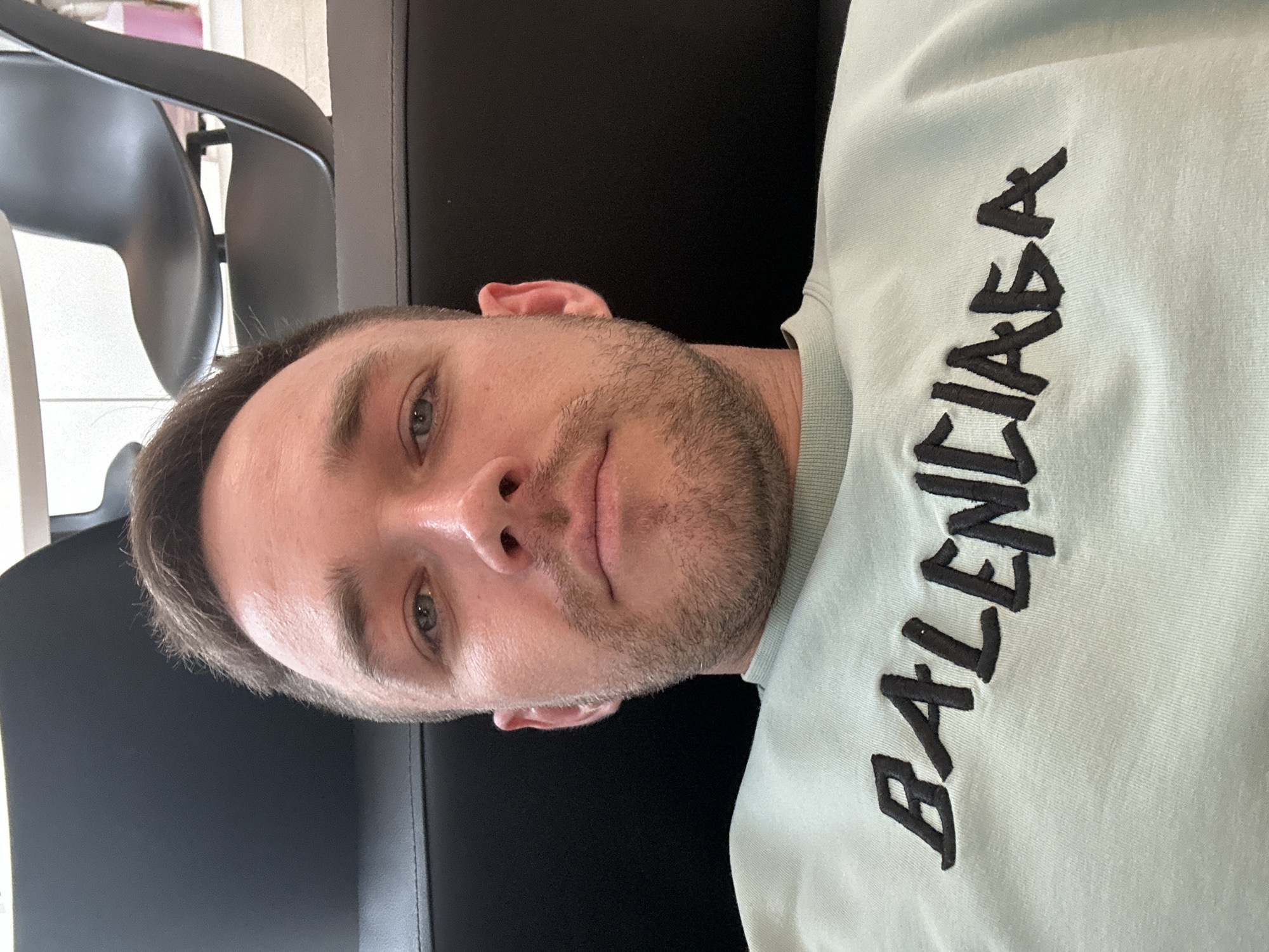 A selfie depicts a man with short, light brown hair, light eyes, and stubble. He's wearing a light green sweatshirt with the word "BALENCIAGA" embroidered in black lettering. He appears to be sitting on a black sofa, with a black chair visible in the background. The location in the city of Perm, Russia, cannot be determined.