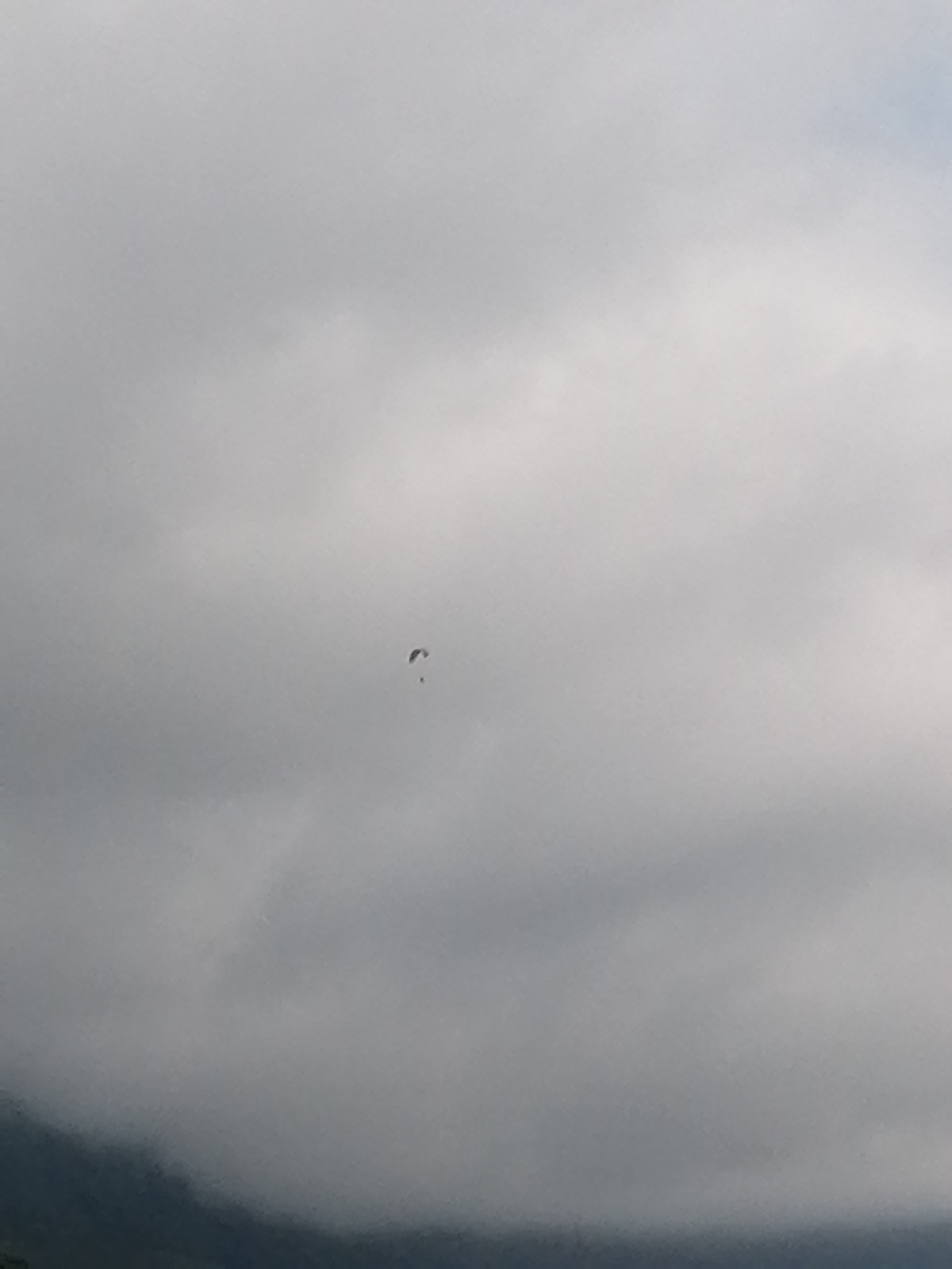 A paraglider is suspended in the sky above Sa Pa Ward, Vietnam. The sky is overcast, with grey clouds filling most of the frame. The paraglider is located near the center of the image and is small relative to the sky. A dark, mountainous or forested area is visible at the bottom of the image.