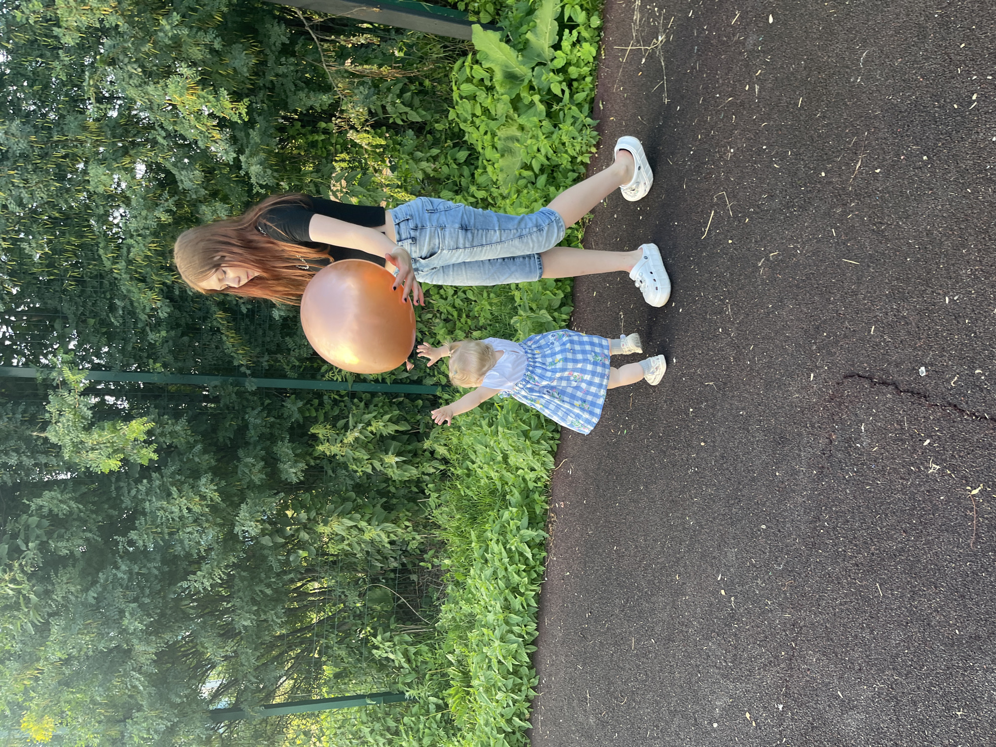 A teenage girl with long red hair is standing on a dark asphalt surface, holding a large, shiny copper-colored balloon. She is wearing a black crop top and light blue capri jeans. Next to her, a toddler in a blue and white checkered dress is reaching up towards the balloon with their left arm. The two are outdoors, with a lush green hedge and trees forming the background, separated by a green metal fence. The lighting suggests it is daytime, possibly late afternoon, with sunlight filtering through the foliage. The asphalt surface appears to be part of a park or recreational area.