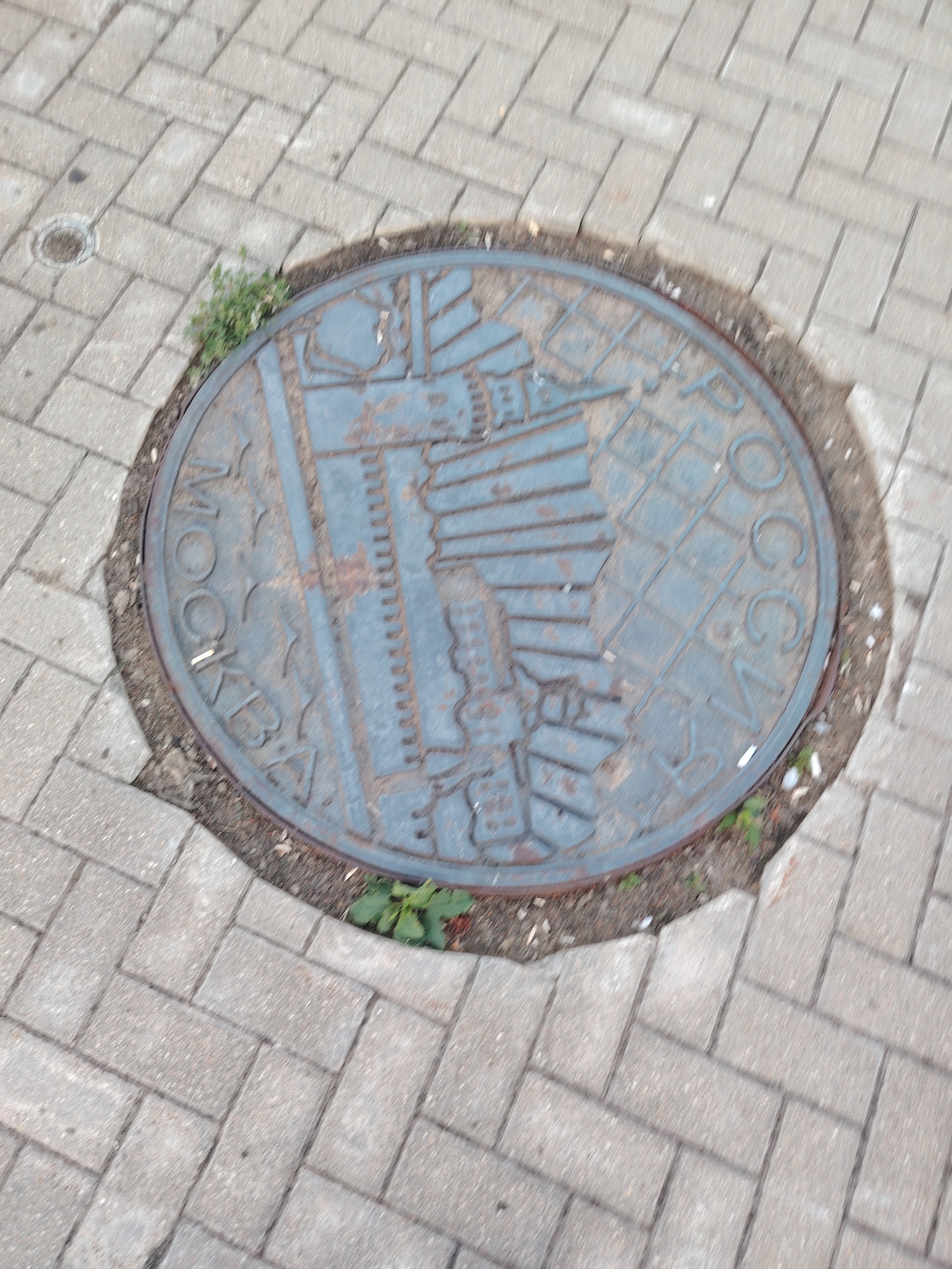 A circular metal manhole cover is embedded in a grey paved walkway. The manhole cover features an embossed design that includes a globe with grid lines and a stylized image of what appears to be a building with several vertical lines, possibly representing windows or architectural features. Cyrillic text is also embossed around the perimeter of the manhole cover. The text on the left side of the cover reads "МОСКВА" (Moskva, meaning Moscow), and the text on the right side reads "РОССИЯ" (Rossiya, meaning Russia). The manhole cover is weathered and shows signs of rust. Some small green weeds are growing around the edges of the manhole cover where it meets the soil. The paved walkway is made of rectangular grey bricks laid in a herringbone pattern. The image is taken from a slightly elevated, oblique angle, causing some motion blur. The overall lighting suggests an outdoor daytime scene. The manhole cover is situated in the city of Konakovo, Russia.