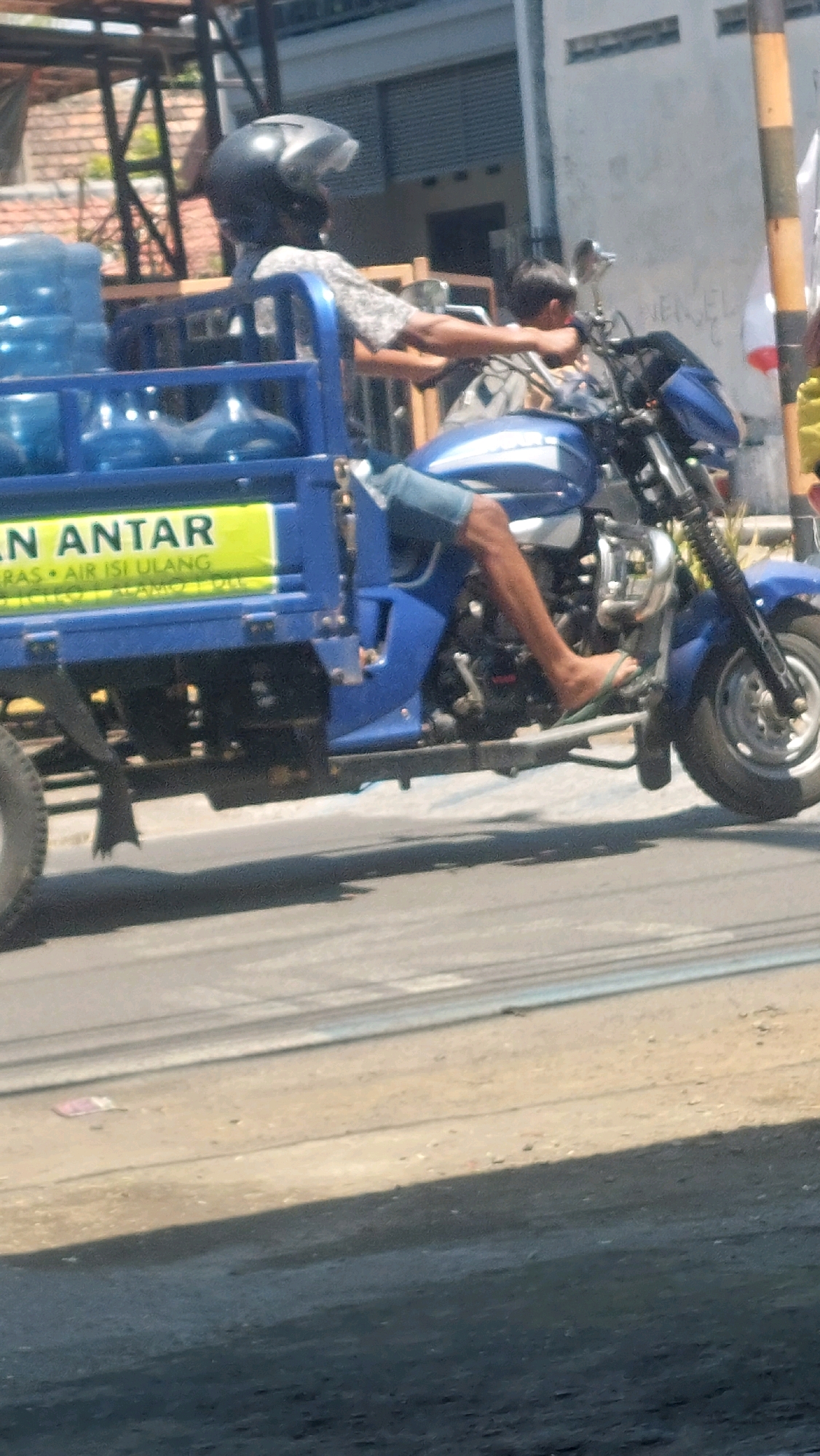 In Sidoarjo, Indonesia, a blue motorized tricycle, commonly known as an "odong-odong," is captured mid-transit. The tricycle is laden with several large blue water containers, suggesting a delivery or refill service. The driver, wearing a helmet and a patterned shirt, operates the vehicle. A second person, a child, is seated behind the driver, facing forward. The tricycle is traveling on a paved road, with the right side of the frame showing a building with white walls. The scene appears to be an everyday occurrence in Sidoarjo, reflecting the city's transportation and commercial activities.