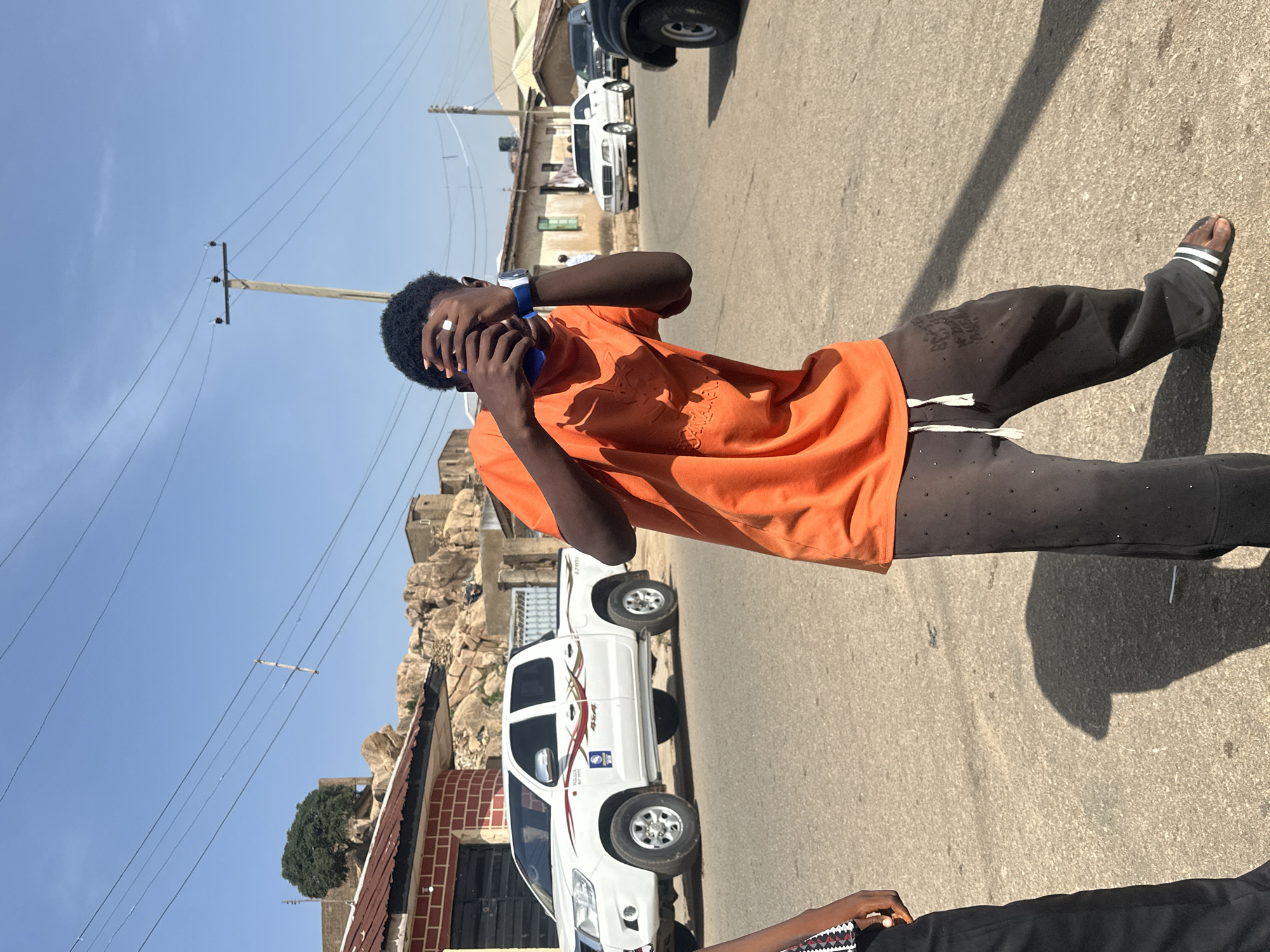 A street scene in Unknown, Unknown is captured from a slightly elevated perspective under clear, bright daylight. The primary subject is an individual of color, wearing a bright orange t-shirt and dark sweatpants with white drawstrings. The person is holding a blue smartphone to their face with their left hand and holding their head with their right hand, as if they are in some distress. They are wearing white and black flip-flops and a ring on their left pinky.
To the left of the individual is a white pickup truck parked along the side of the road and an unidentified building. Behind the white truck is a large rock formation. A power pole is visible extending into the blue sky, with power lines stretching across the frame. Other vehicles are visible in the distance, further up the road. The pavement shows signs of wear and tear. Another individual's legs are visible at the bottom of the frame, dressed in black.