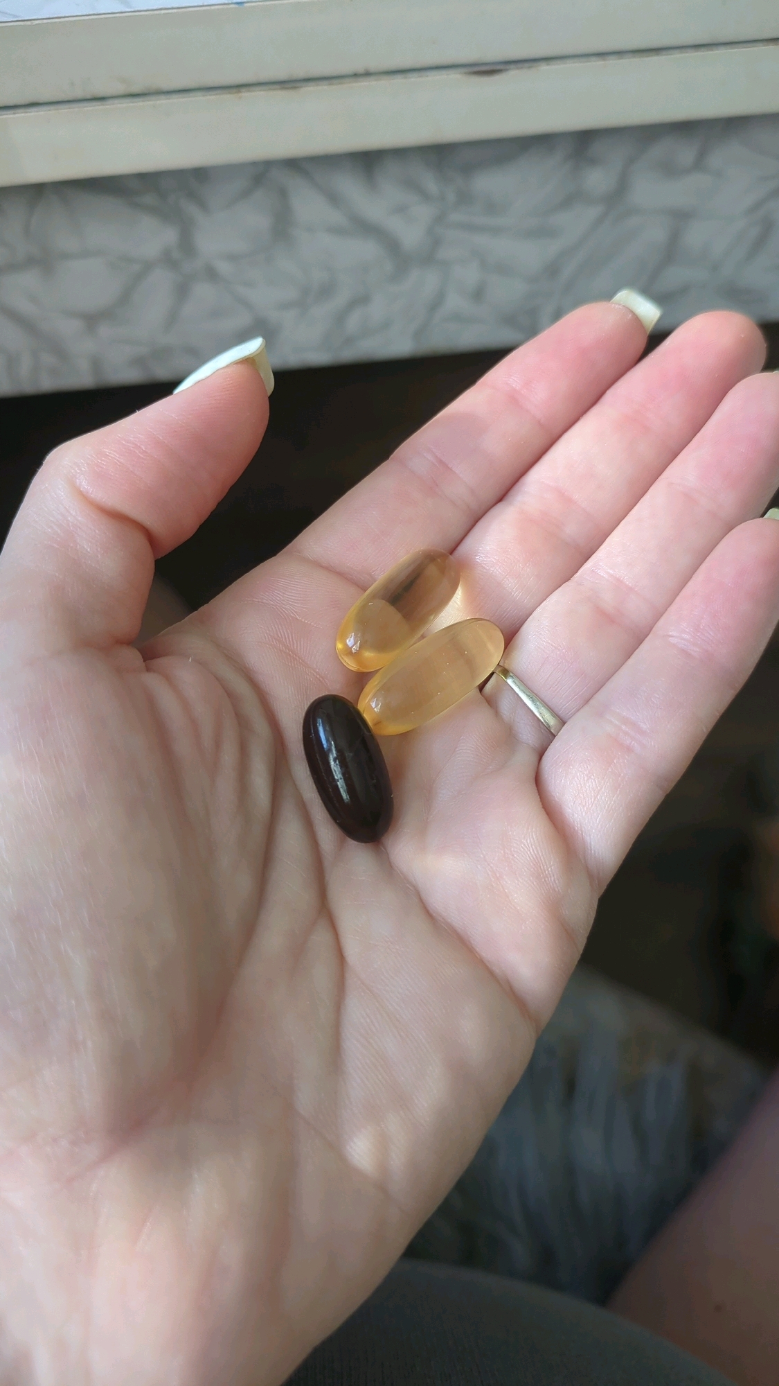 A person's hand, with pale skin and light-colored fingernails, is shown from a close-up perspective. The hand is open, palm up, and contains three oval-shaped pills. Two of the pills are translucent and yellow, while one is opaque and dark brown. A silver ring is visible on the ring finger of the hand. The background is out of focus, but appears to be an indoor setting with a patterned surface behind the hand. There is no identifiable information in the image to suggest a location in Chișinău, Moldova.
