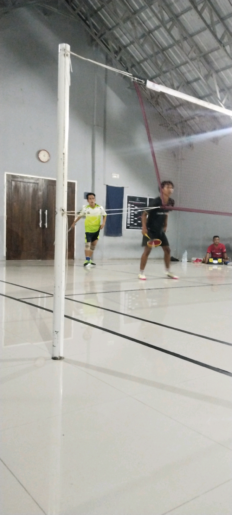 In the Kliris, Indonesia city, this image captures a badminton match in an indoor court. Two young men are actively playing, one in a neon green and white shirt and black shorts, the other in a black shirt and shorts. A third man in a red shirt is seated on the floor in the background. The court features a white tiled floor with black lines. A white pole with a net divides the court. The ceiling appears to be made of corrugated metal.