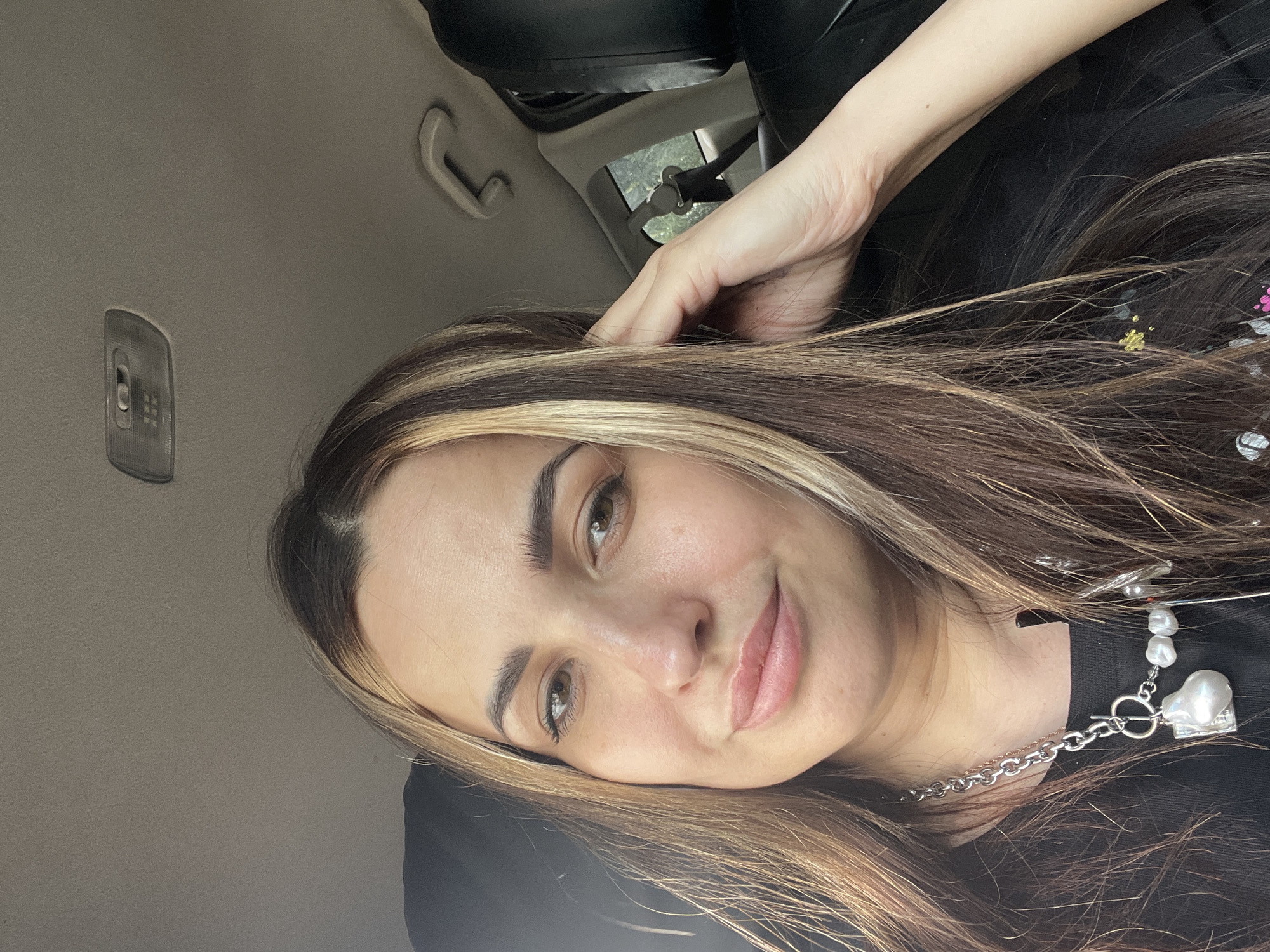 A young woman with long brown hair with blonde highlights is sitting in what appears to be a car. She is smiling and looking directly at the camera, with her right hand resting on the side of her head. She is wearing a black top and a silver necklace with pearl embellishments. The interior of the car is visible, including the ceiling, a car light, and a seatbelt. The location is Unknown, Unknown.