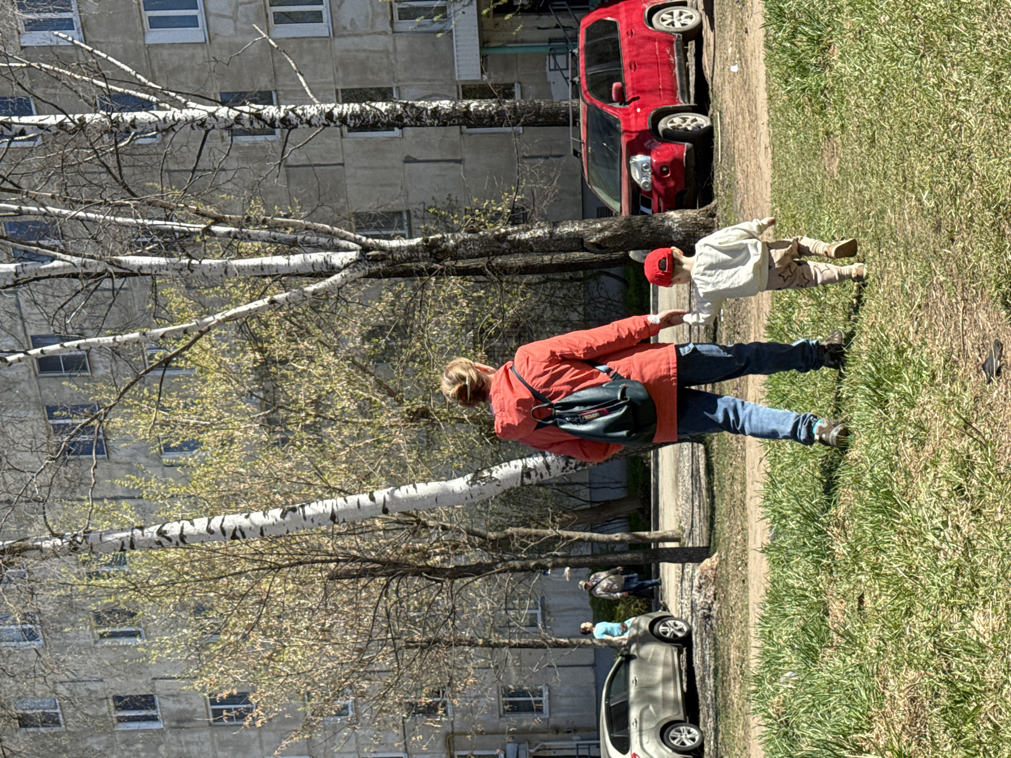 The image shows a woman and a child walking on a grassy area next to a building in Izhevsk, Russia. The woman is wearing a red jacket, blue jeans, and a black backpack. The child is wearing a red hat, a light-colored shirt, and patterned pants. They are holding hands. Behind them are parked cars, including a red SUV and a light-colored sedan. Several birch trees are visible in the foreground, with bare branches and some new leaves. The building in the background is a multi-story apartment complex with rows of windows. Two additional people can be seen distantly walking in the same direction as the woman and the child.