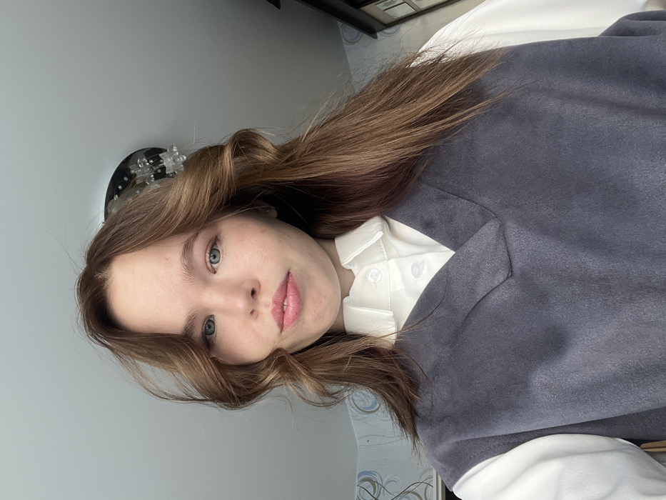 An indoor self-portrait photograph captures a young adult female from the chest up, looking directly at the camera. The subject has long, wavy brown hair, blue-green eyes, and light pink lips, displaying a neutral facial expression. She is dressed in a white collared button-up shirt worn under a dark grey, V-neck, sleeveless top. The background features a light-colored ceiling, with a dark-based, clear-element ceiling light fixture partially visible in the upper right quadrant. A light-colored wall with a subtle blue and grey patterned wallpaper is discernible on the left, while a dark piece of furniture is partially visible on the far right. The scene is located within a building in the city of Chaikovsky, Russia.