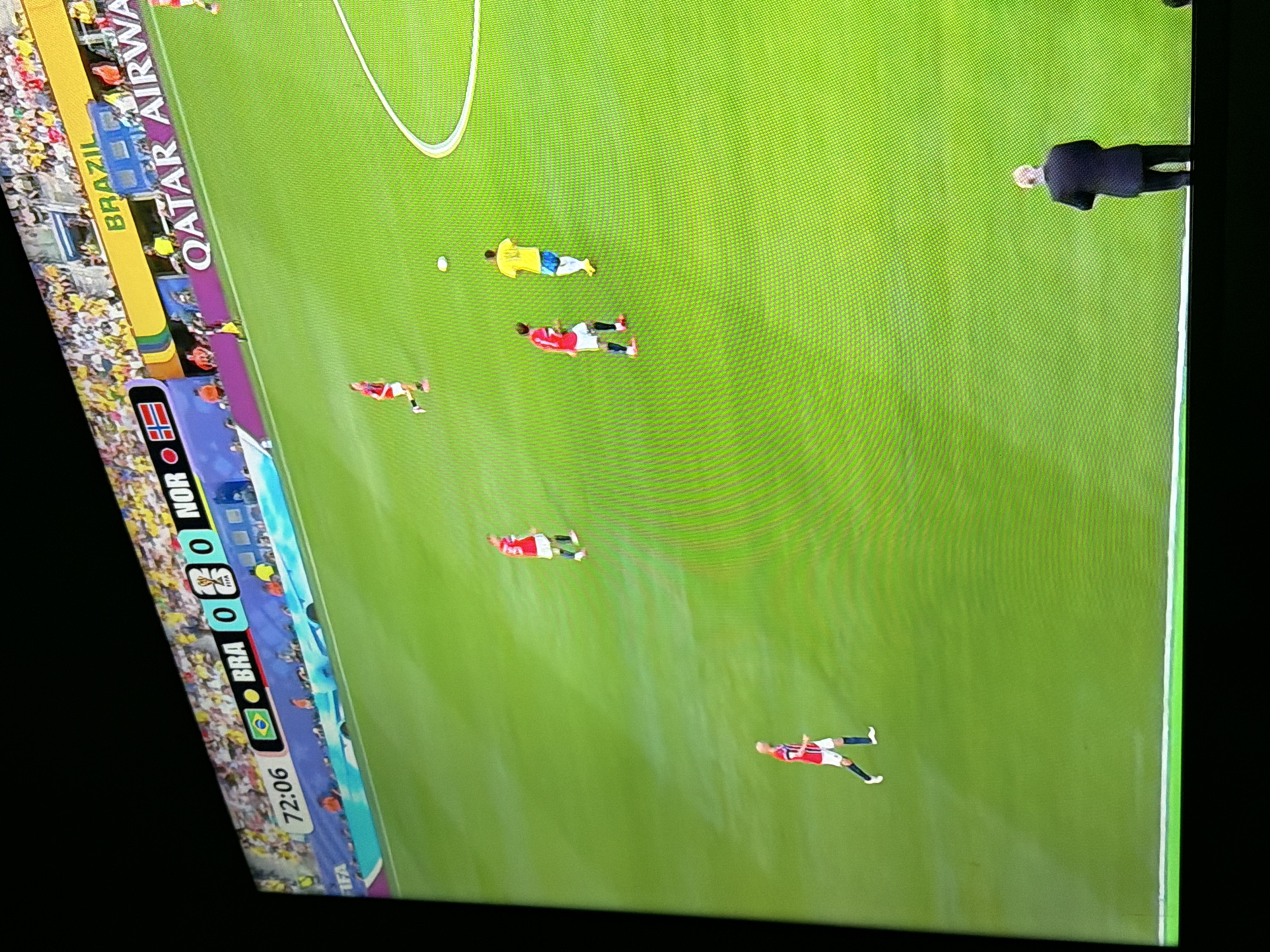 The image shows a soccer match on a bright green field. The scoreboard indicates that the game is between BRA (Brazil) and NOR (Norway), with the score currently 0-0 in the 72nd minute. Several players in red and white uniforms and one player in a yellow uniform are visible on the field. A man in a dark suit stands near the bottom right corner of the frame, facing away from the camera. In the background, spectators are visible in the stands, many wearing yellow, suggesting support for Brazil. Advertising boards for "QATAR AIRWAYS" are also present. The image does not provide any information about the city of Ognianovo, Bulgaria.