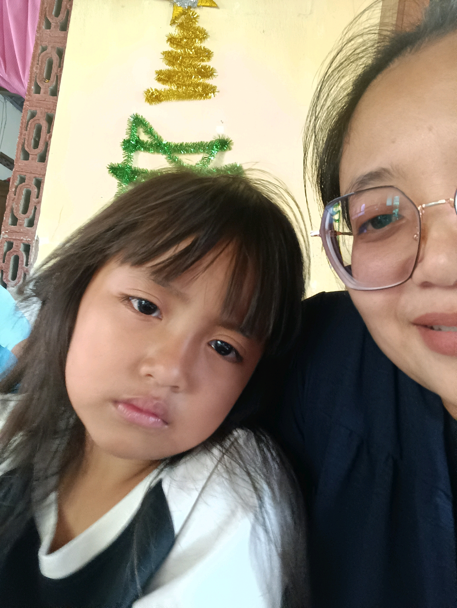 The image is a close-up, selfie-style shot of two individuals. On the left is a young girl with dark hair and bangs, looking directly at the camera with a neutral expression. She is wearing a white shirt with black sleeves. To the right is an adult, likely a woman, wearing glasses with gold frames and a dark top. Her right eye and part of her cheek are visible, and she appears to be smiling slightly.
In the background, a wall is painted a light yellow color. There are two Christmas-themed decorations on the wall: a gold tinsel Christmas tree with a silver star at the top, and a green tinsel garland looped below it. To the far left, a section of a decorative brick wall is visible, with a pink curtain behind it. The presence of Christmas decorations suggests the photo was taken during the holiday season. The metadata of the image indicates the location as Ploso, Indonesia.
