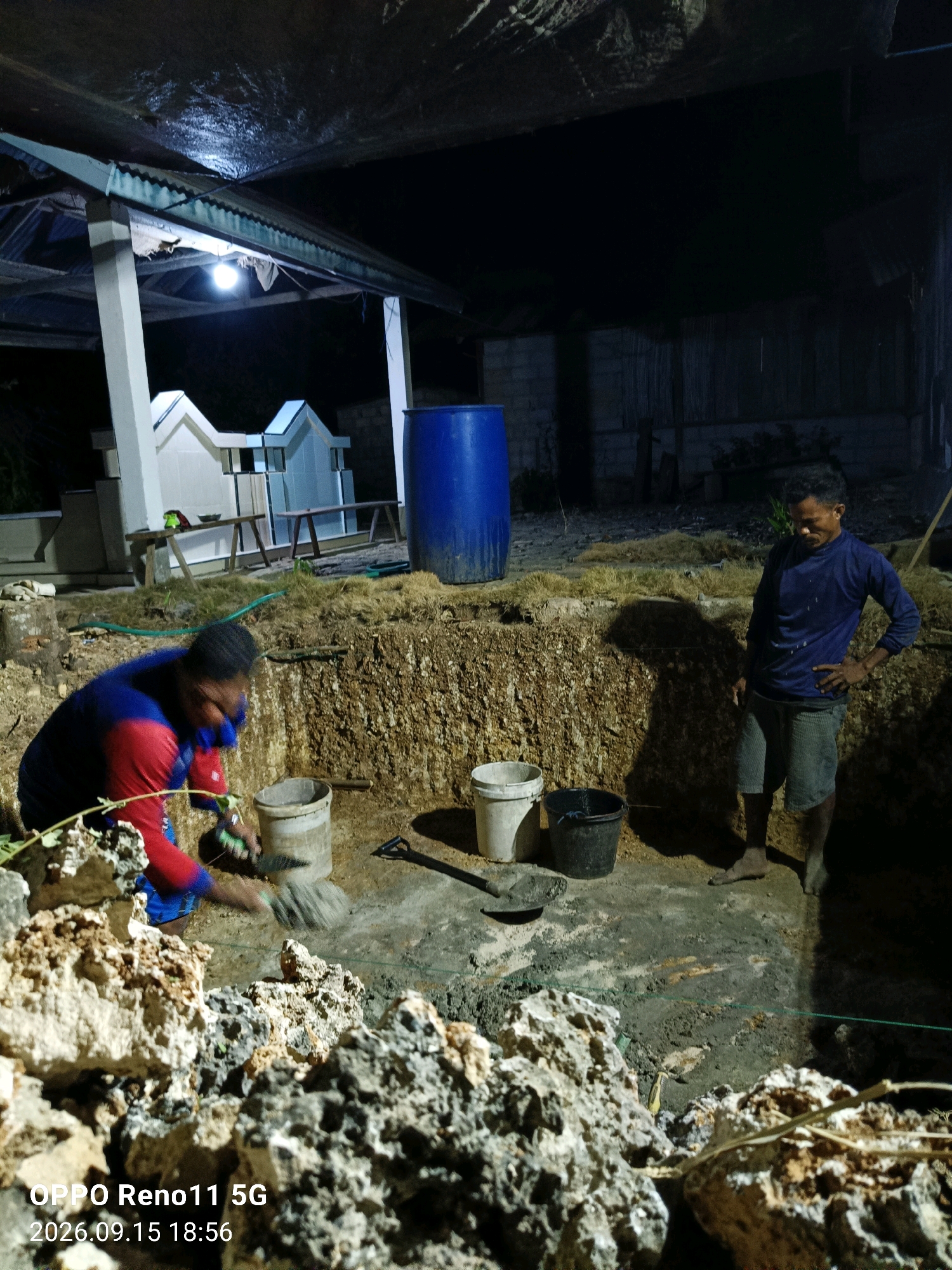 The image depicts an outdoor construction site at night, possibly in Otan, Indonesia, characterized by dim lighting from an overhead bulb. Two individuals are present. In the foreground, a person wearing a blue and red long-sleeved shirt and blue shorts is actively working, seemingly digging or mixing materials with a tool and a bucket. Their face is partially obscured. In the midground, another person in a blue long-sleeved shirt and gray shorts stands with one hand on their hip, observing the work. They appear to be bare-footed.
The immediate environment is a deep pit with rough, earthy walls, suggesting excavation work. Several buckets, one white and one black, are positioned near the pit. A shovel lies on the ground. In the background, a large blue barrel stands prominently. Further back, a covered structure with white pillars and a corrugated roof is visible, and behind that, a darker, less defined area with what appears to be a wooden fence. Rocks and dry vegetation are scattered around the site. The ground is uneven and appears to be a mix of soil and some construction debris. The overall impression is of manual labor taking place during nighttime hours.