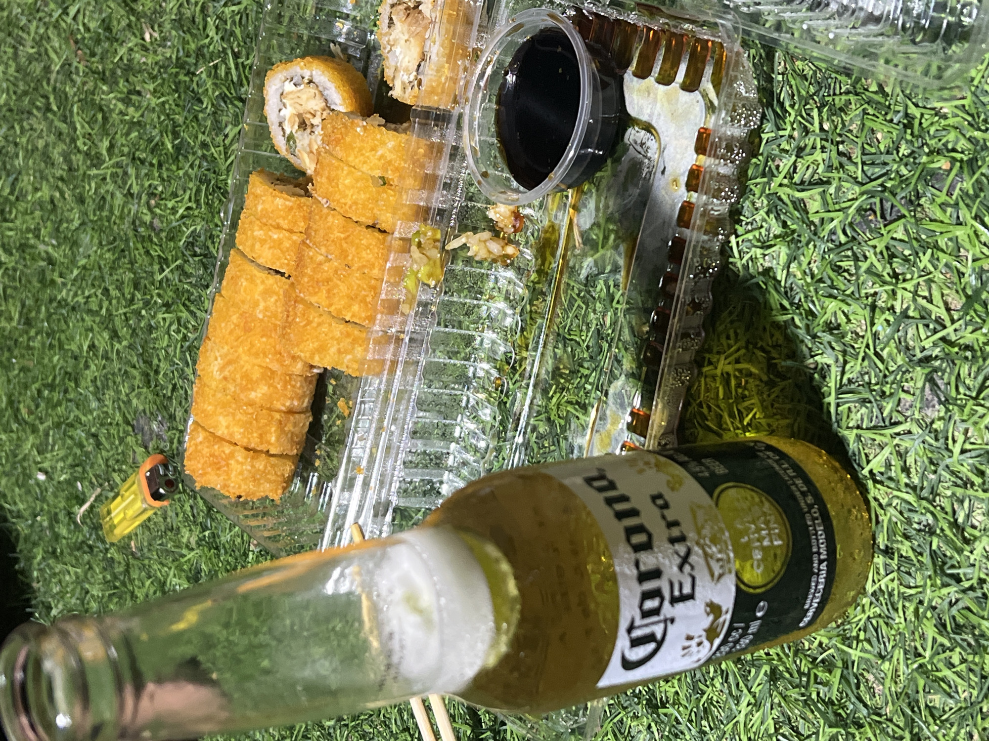 The image shows a plastic container of fried sushi rolls, a small container of dark sauce, a yellow lighter, and a bottle of Corona beer on a patch of artificial green grass. The sushi is cut into bite-sized pieces. The scene appears to be outdoors, possibly on a patio or deck covered with artificial grass. It is not possible to confirm the location in the city of Arica, Chile.