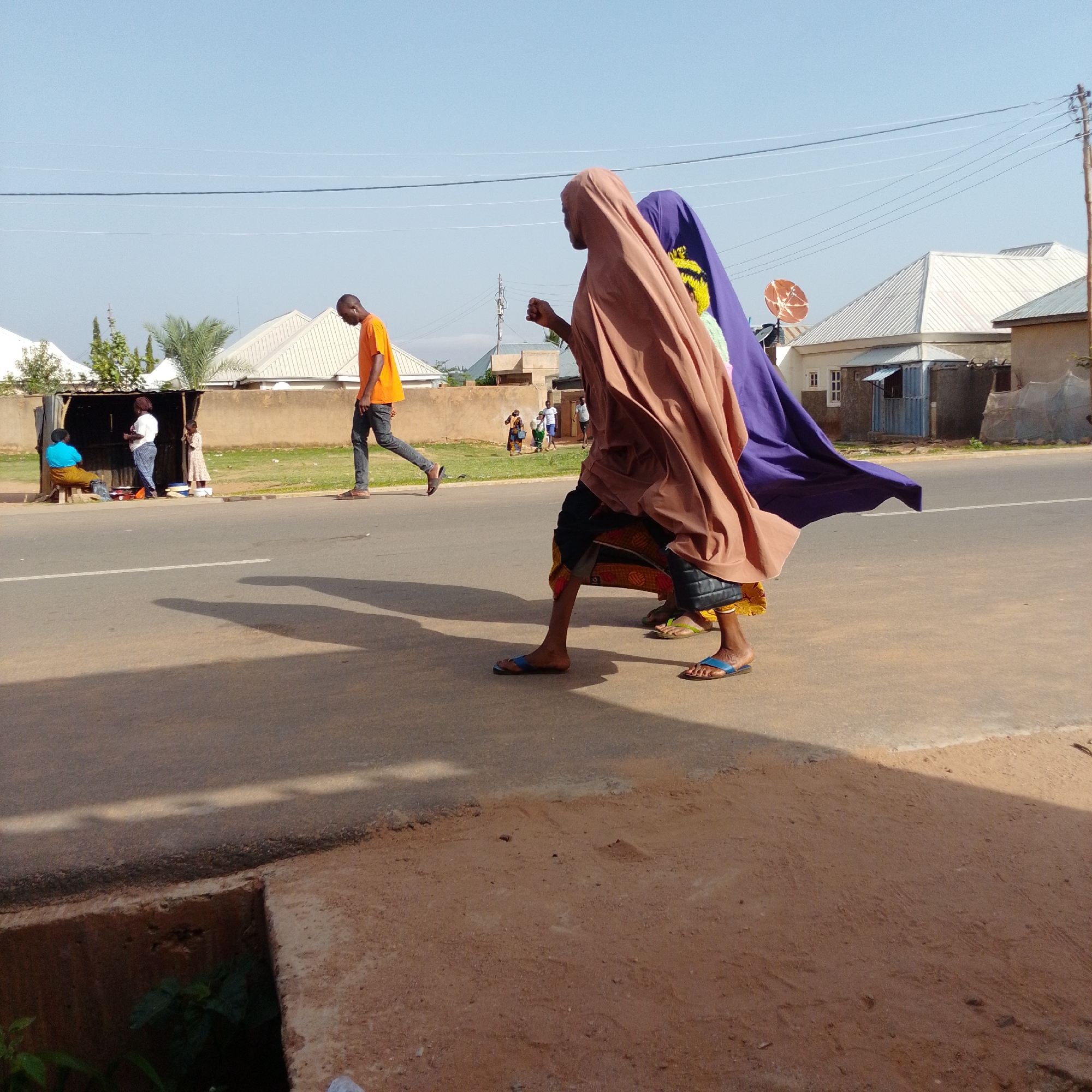 In the city of Unknown, on a bright, clear day, two young women, dressed in traditional Islamic attire, walk on the right side of a paved road. The woman on the left wears a brown hijab and a dark skirt, while the woman on the right is adorned in a purple hijab and a yellow and green patterned skirt. They are walking side-by-side, with their legs and feet visible in the foreground. Both are wearing sandals. To the left of the women, a man in an orange t-shirt and dark pants walks in the opposite direction, his gaze lowered. Further to the left, a small outdoor market stall is visible with three people interacting. A woman in a blue top and yellow skirt sits on a bench, a woman in a white top and patterned pants stands, and a young girl in a patterned dress stands nearby. The market stall has a corrugated metal roof and is open to the street. The road has a white line marking its center and is bordered by a dirt path and grassy area on the left. In the background, residential buildings with white roofs and satellite dishes are scattered across the landscape under a clear blue sky with thin, high-altitude clouds. Telephone poles and wires run across the upper portion of the image. The overall scene depicts a typical day in a community where traditional and modern elements coexist.