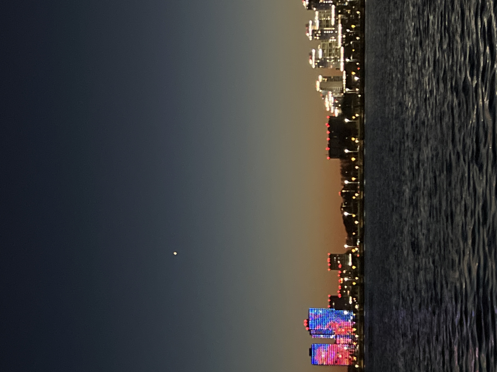 Here's a detailed description of the image:
**General Description:**
The image depicts a cityscape at night reflected on a body of water. The perspective is somewhat unusual, as the image seems to be rotated ninety degrees clockwise.
**Environment:**
* **Sky:** Dark gradient, transitioning from a deep blue/black at the top to a lighter tone, likely indicating a dusk or dawn atmosphere. A small bright dot is visible in the dark sky, possibly a star or planet.
* **Water:** A body of water takes up the right side of the image. Its surface is textured with ripples and waves that reflect the city lights.
* **Cityscape:** A skyline of buildings, illuminated at night, runs along the line where the sky meets the water.
**Objects and Subjects:**
* **Buildings:** Various buildings of different heights and architectural styles. Some are illuminated with white lights, while others have colored lights (red, blue, and purple are visible). Some of the lower buildings have red lights on top of them, which is common for taller buildings.
* **Lights:** A significant number of streetlights or other ground-level lights are interspersed along the skyline.
* **Reflection:** The reflections of the buildings and lights are visible on the water surface.
**Action and Interactions:**
* The lights from the city are reflecting off the water.
* There are no discernible people or specific actions taking place in the image, aside from the implied human presence of a city at night.
**Location Details:**
* The image depicts the city of Kazan, Russia, as evidenced by the unique lighting and architecture visible in the buildings.
