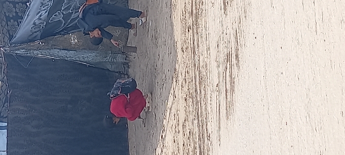 The scene depicts two individuals on sandy, uneven ground in Khan Yunis, Palestinian Territory, against a backdrop of makeshift structures. A person, identified as a child or young individual, is squatting on the left, wearing a red long-sleeved top, dark trousers, and light sandals, with a dark backpack slung over their shoulders. This individual is focused downwards towards the ground. To the right, another child or young individual, dressed in dark clothing and light-colored sandals, bends forward at the waist. This individual holds a cylindrical metal container in their left hand and points towards the ground with their right index finger. A light-colored bag is visible over their right shoulder. Behind the individuals are temporary shelters or walls constructed from various materials, including dark tarpaulin and fabric with faint floral patterns. The ground in the foreground shows horizontal striations.