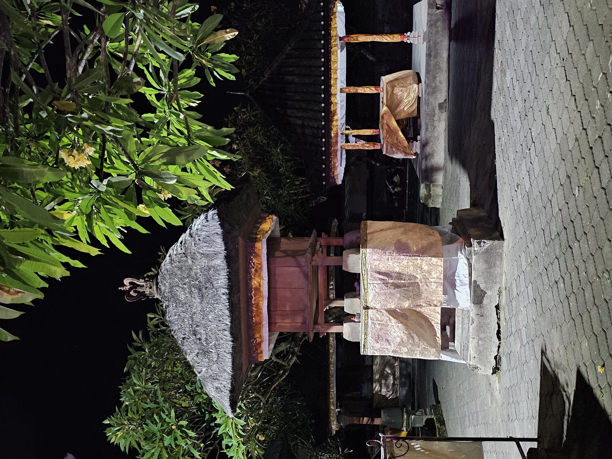 The image depicts an outdoor scene at night, likely a temple complex in Denpasar, Indonesia. In the foreground, a large, multi-tiered structure with a thatched roof is visible. This structure has a reddish-brown wooden body supported by pillars, and is adorned with ornate golden and white fabric draping its base. A small, metallic crown-like ornament sits atop the thatched roof. To the right, a smaller, open-air structure with a dark tiled roof is also present, similarly decorated with golden fabric and supported by pillars. Both structures are situated on a paved courtyard made of interlocking hexagonal stones. Lush green foliage from overhanging trees, some with small yellow flowers, frames the upper portion of the image, contrasting with the dark night sky. The lighting suggests artificial illumination, casting shadows on the paved ground. No people are present in the scene.