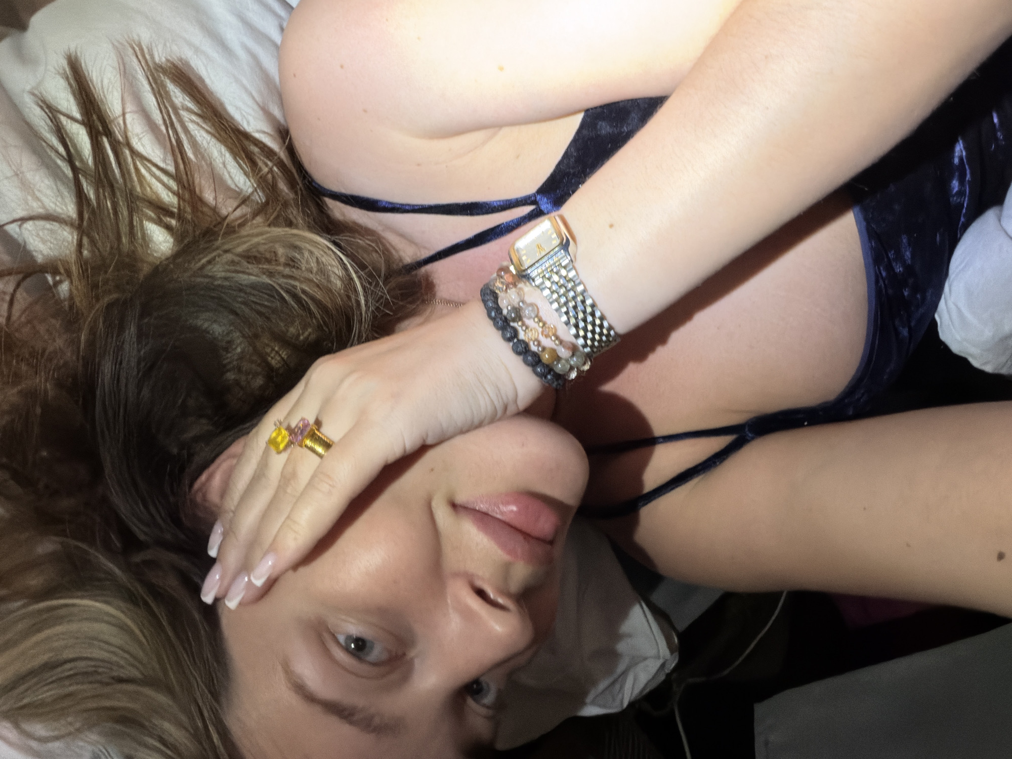A woman lies on a bed in an indoor setting. She is wearing a blue top with thin straps and several bracelets on her right wrist along with a watch. She also wears a large gold ring with colorful stones on her left hand. The location is Высокая Гора, Russia.