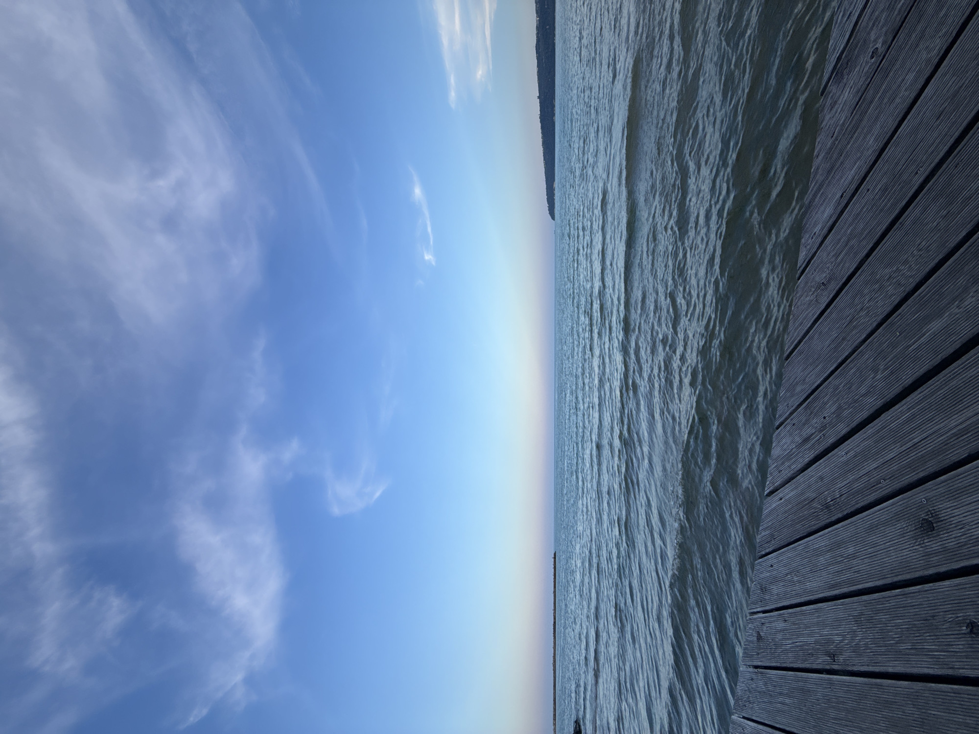 The image depicts a scenic view of the sea with a wooden pier in the foreground. The sky is a gradient of blue, transitioning from a deeper hue at the top to lighter shades near the horizon, with wispy white clouds scattered across it. The sea is calm with gentle ripples, reflecting the sky's colors. In the distance, a man-made structure, possibly a breakwater or pier, extends into the water on the left side of the image. On the right side, a landmass with trees and buildings is visible, suggesting a coastal city or town. The wooden pier in the foreground is made of dark planks laid horizontally, with visible grain and texture, and it leads the eye towards the expansive sea and sky. The overall atmosphere is serene and peaceful. This scene is likely located at the Varna Sea Garden or along the Varna coastline in Varna, Bulgaria, given the characteristic landscape of the Black Sea coast. There are no people or animals visible in the image.