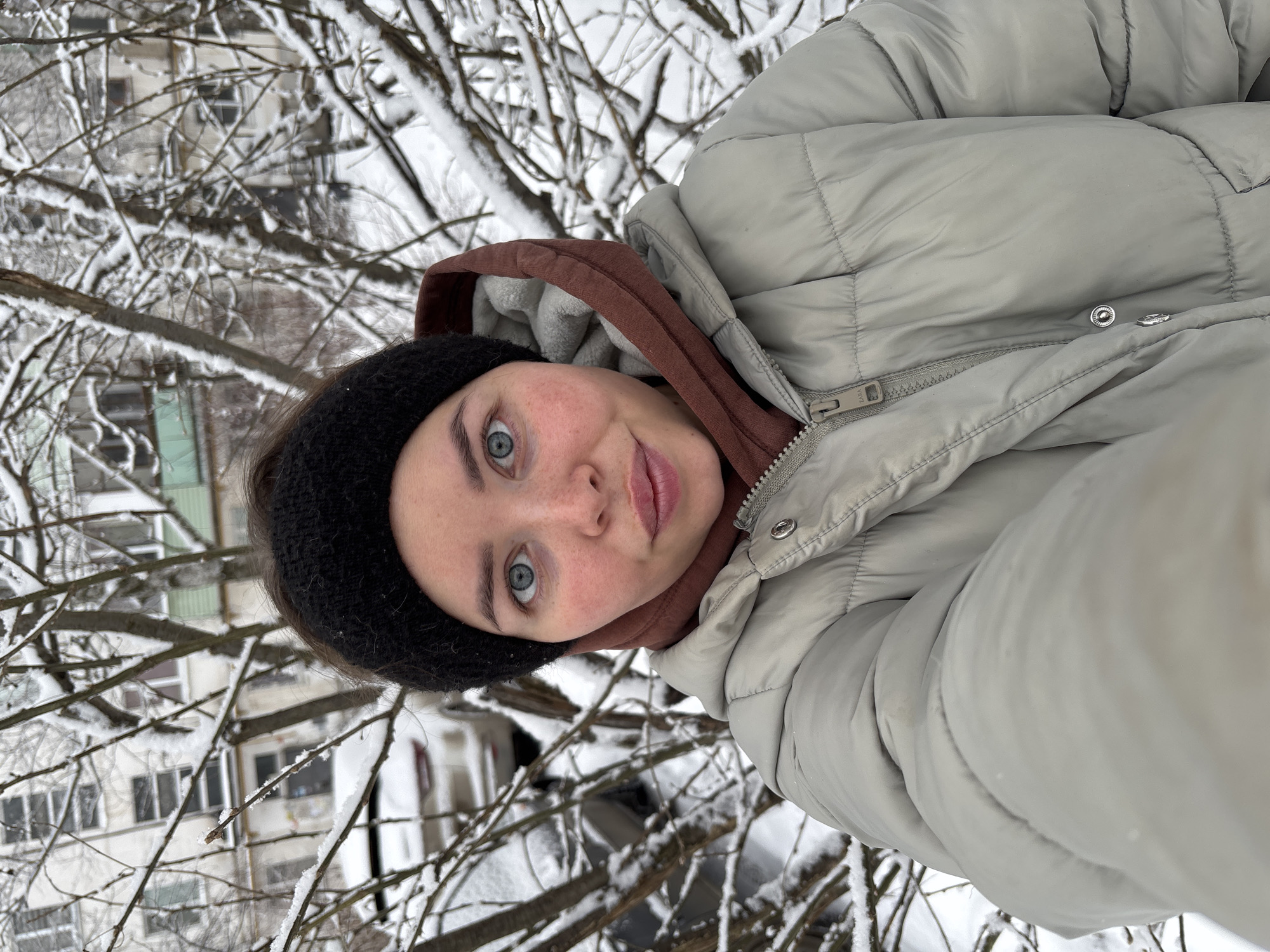 A woman is taking a selfie outdoors in winter, presumably in Izhevsk, Russia, based on environmental cues. She is wearing a light gray puffer jacket over a brown hooded sweatshirt and a black headband. Snow-covered branches of trees are visible in the background, along with residential buildings suggesting an urban setting.