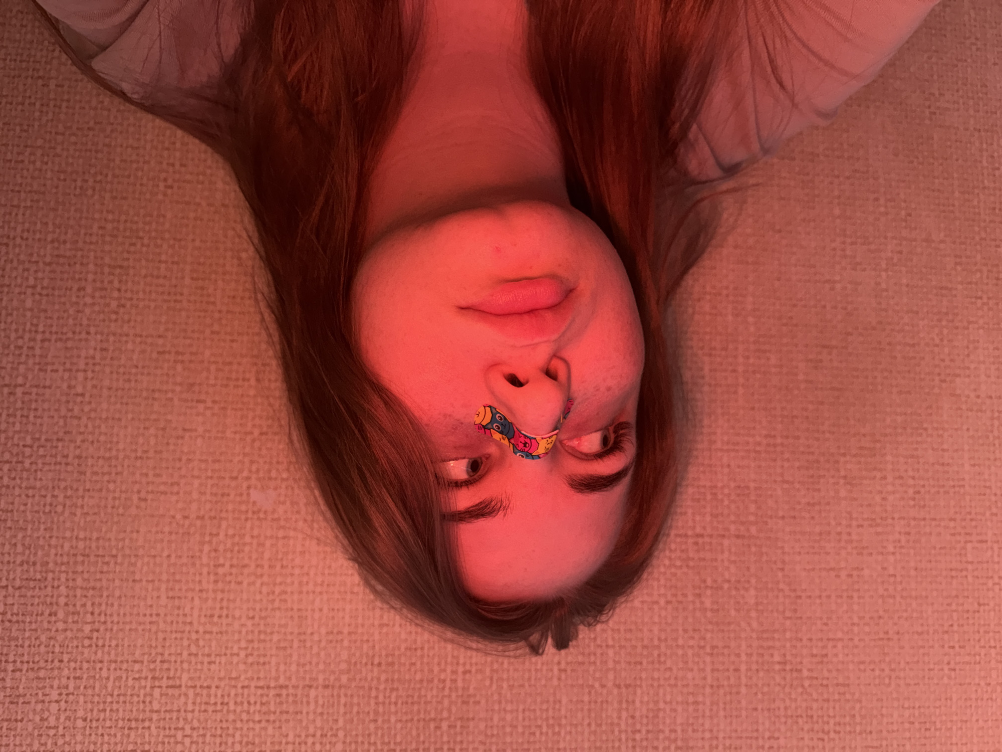 A person with fair skin and auburn hair, illuminated by a red light, is lying down, possibly on a textured surface. A colorful bandage covers the bridge of their nose. The person's eyes are open, looking slightly upward. The location, Chaikovsky, Russia, is not discernible from the visual information.