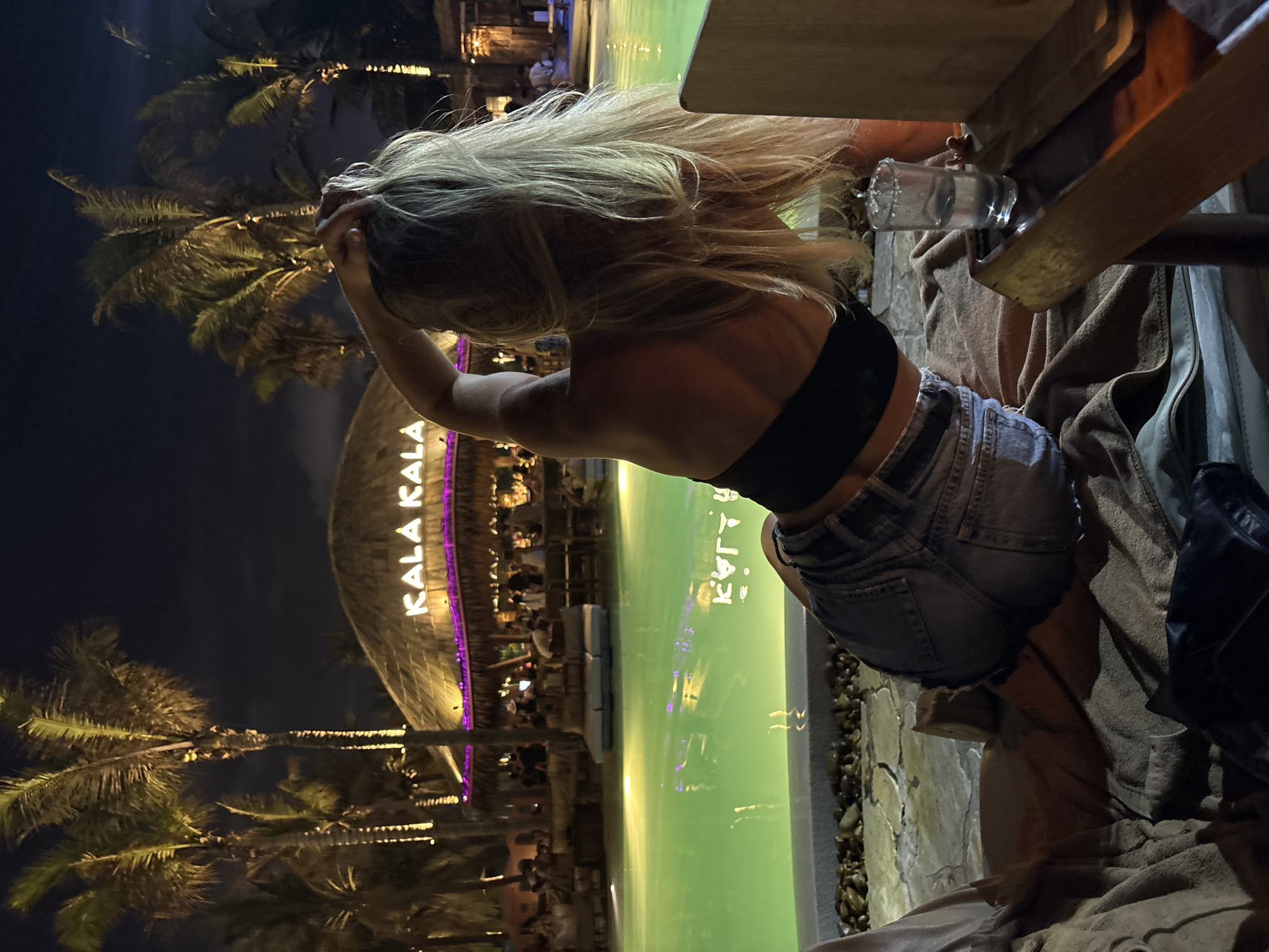 At night, a young woman is at a bar at the Kala Kala Beach Club in Đà Nẵng, Vietnam. She has blonde hair, a black top, and denim shorts. Her hands are near her head. Palm trees are visible in the background. There is a building with “KALA KALA” written on it in pink. A bright light emanates from the building and reflects on a pool. The young woman is sitting at a table with a drink. She is sitting on a blanket or towel.