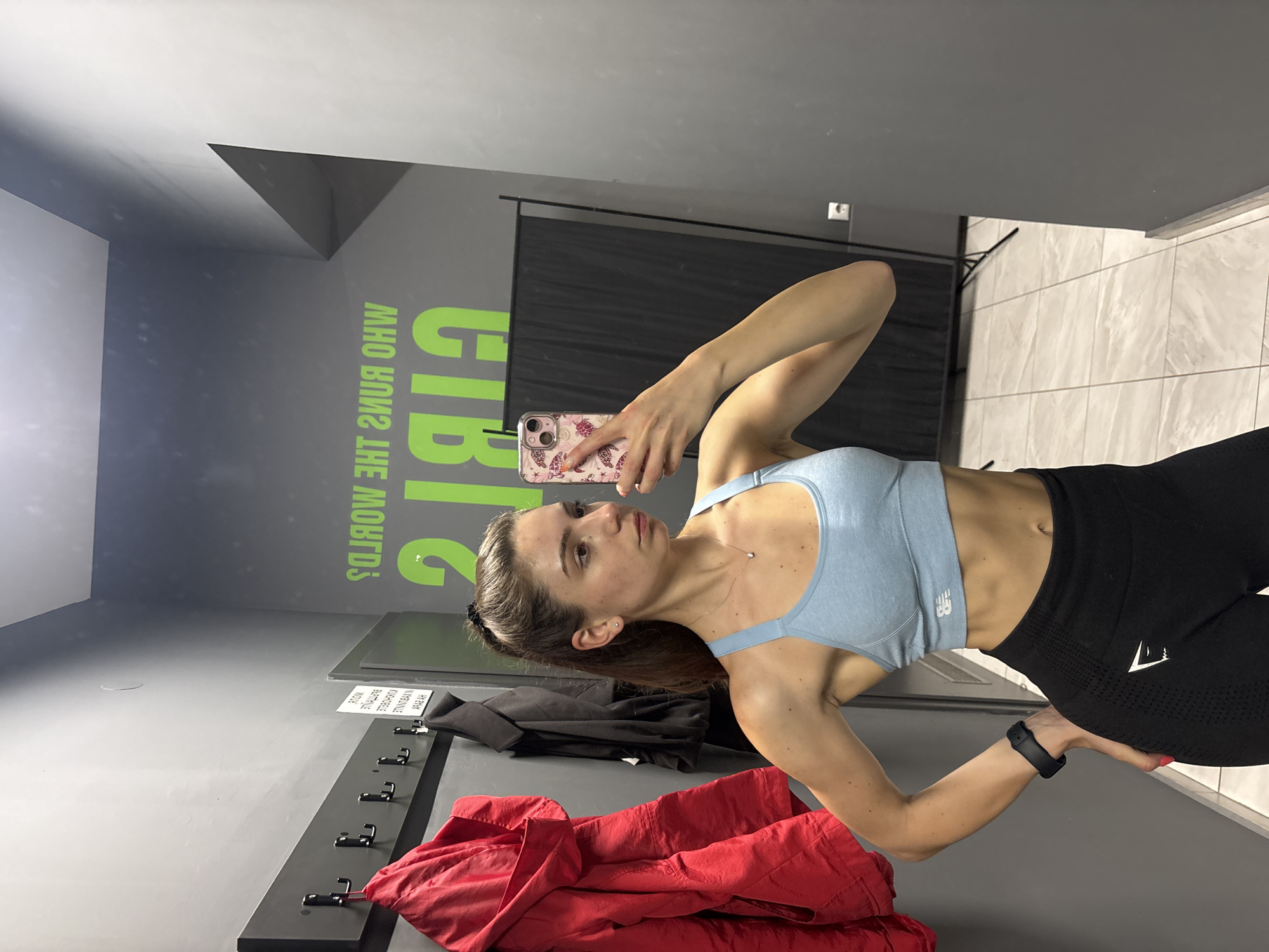 A woman takes a selfie in a locker room. She wears a light blue sports bra and black leggings. The locker room has a gray wall with hooks, and a red jacket is draped over the hooks. On the wall is the phrase "Girls Who Run the World." A backdrop is also present. The location in the city of Varna, Bulgaria is unknown.