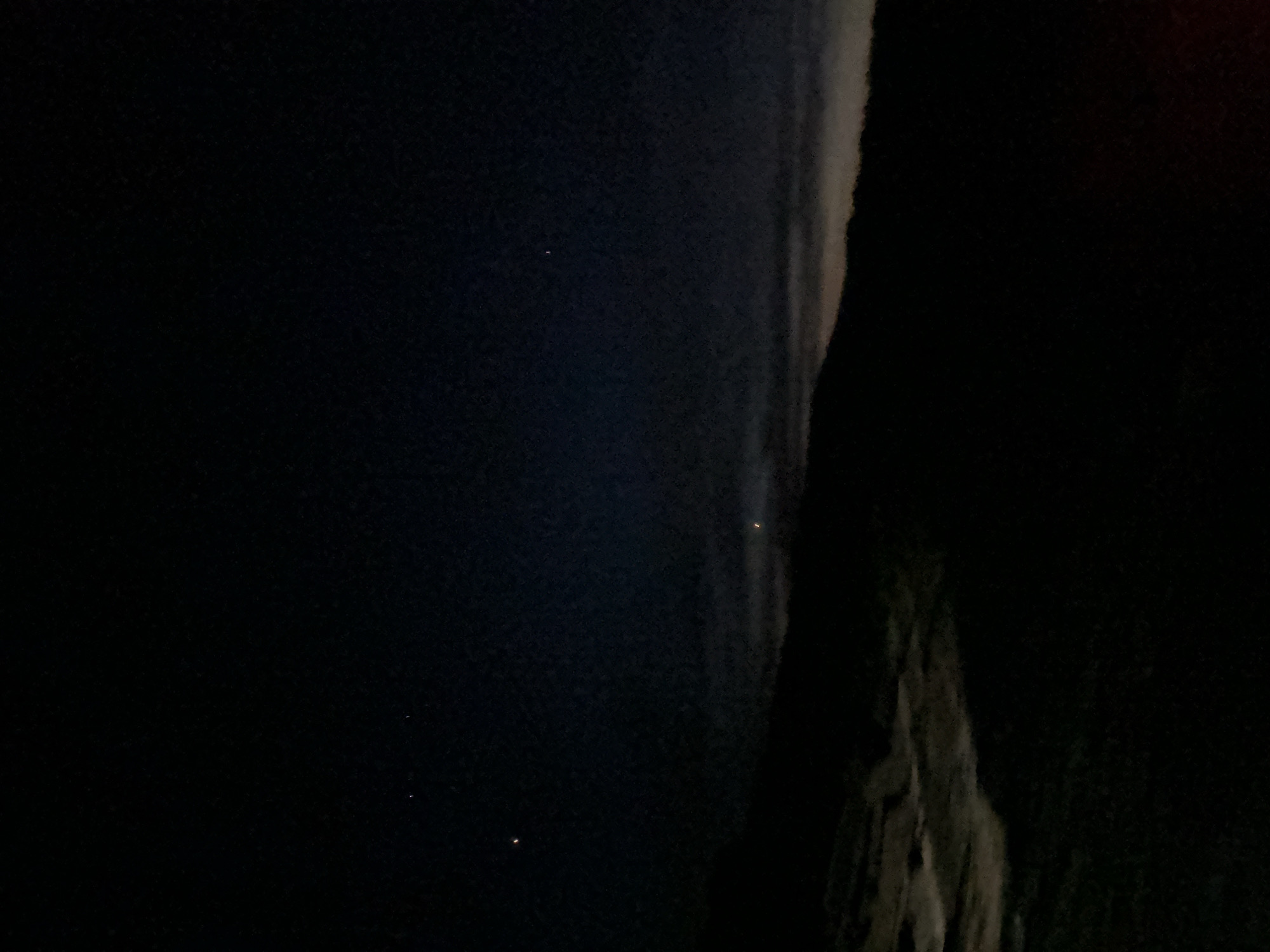 The image depicts a dark night scene in an unknown location. The sky is a deep black with a few faint, distant lights. To the right is a dark, indistinct mass, possibly a rock formation or building, partially illuminated with a faint, irregular pattern. It is impossible to determine the exact location or any other details about the scene from this image.