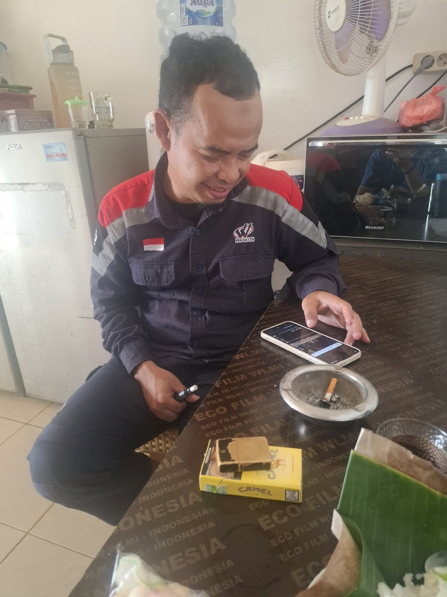 In an indoor setting, likely a breakroom or common area in Ciheulang Tonggoh, Indonesia, a man wearing a dark blue work shirt with red and grey accents is seated at a table. The shirt has a "WASKITA" logo and an Indonesian flag patch. He is looking down at a smartphone in his right hand, which rests on a dark brown table. Next to the phone is a silver ashtray containing a lit cigarette. A yellow pack of Camel cigarettes and a gold-colored lighter are also on the table. The table surface is covered with a material printed with repeating text, including "INDONESIA" and "ECO FILM WLMI". In the background, a refrigerator with "AQUA" branding is visible, along with a white electric fan and a dark appliance labeled "SHARP". Other objects on shelves include a large water bottle and glassware. The overall impression is of a casual break moment for a worker.