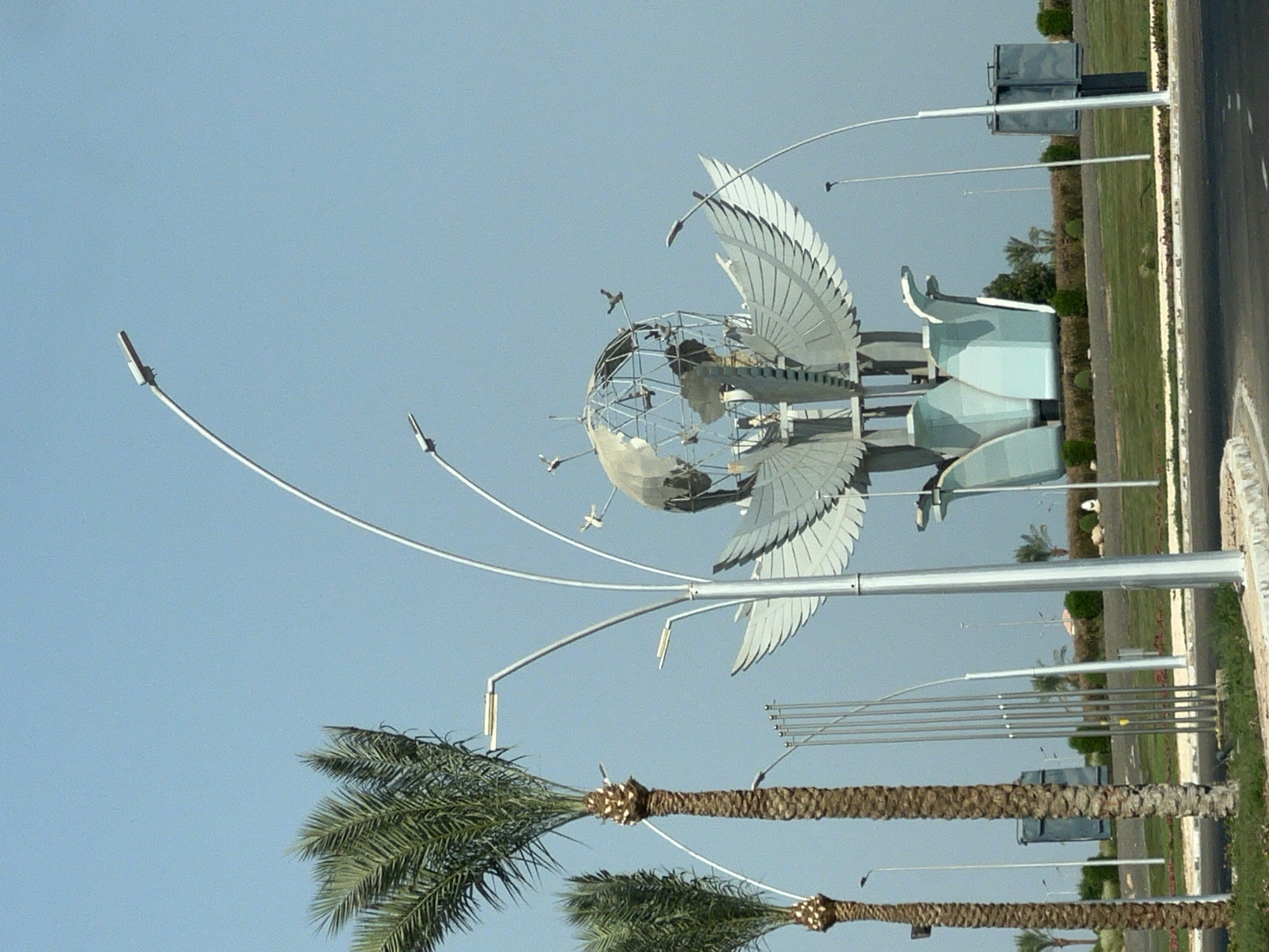 This is an outdoor, daytime, eye-level shot of a public monument in Sharm El Sheikh, Egypt. The monument appears to be a large, stylized sculpture of a globe with wings, positioned in a median or park area. The globe is a metallic geodesic dome, and the wings are also metallic, fanning out from the base. Below the wings are several light blue, petal-like structures. The monument is surrounded by a landscaped area with grass and bushes, and a road is visible in the foreground. Several tall, modern streetlights with curved arms are also present, some framing the monument. Palm trees, characteristic of the region, are scattered throughout the scene, with two prominent ones to the left of the monument. In the background, there are more trees, some low buildings, and a clear, light blue sky. There are no people visible in the image. The overall impression is one of a landmark in a warm, sunny climate.