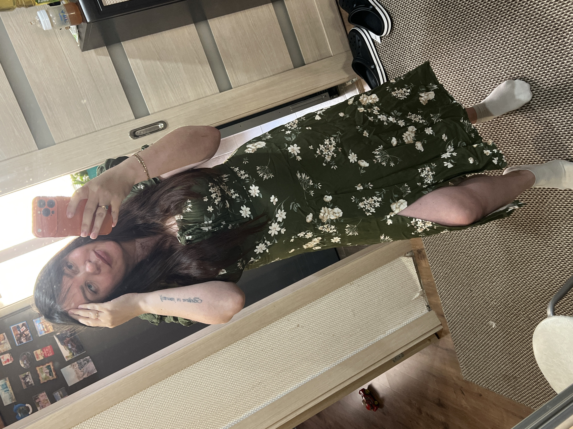A woman is taking a mirror selfie in an indoor setting. She is wearing a green floral print dress with a thigh-high slit and white socks. She is holding her phone in her right hand and touching her hair with her left. In the background, there is a doorway, a mirror, a rug, a pair of black clogs, a small red toy car, and a cabinet with a baby bottle on top. The location is identified as Noviy, Russia.