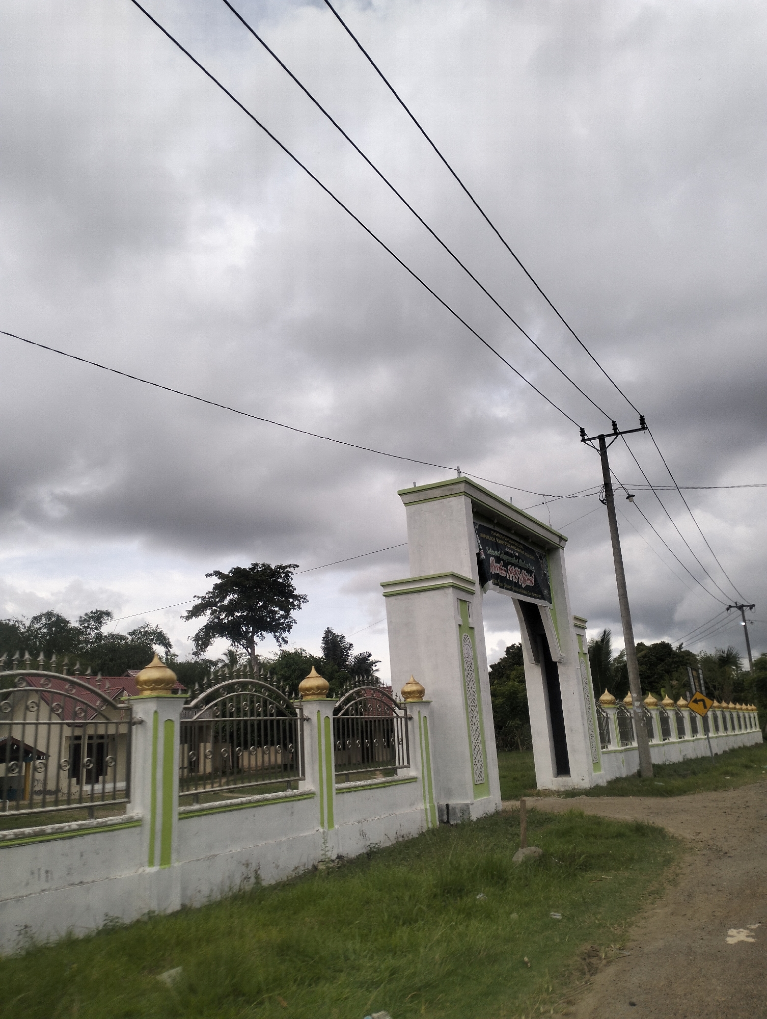 This image shows an outdoor scene with a cloudy sky. In the foreground, there is a white gate with green accents, decorated with intricate patterns. Beyond the gate, a metal fence with decorative golden domes runs along the property line. Behind the fence, several buildings with red roofs are visible, partially obscured by trees. A dirt road curves to the right of the gate and fence. A utility pole with power lines stretches across the upper portion of the image. The scene appears to be located in a rural or suburban area, possibly a religious institution, given the architecture. The location is identified as Payadua, Indonesia. There are no people visible in the image.
