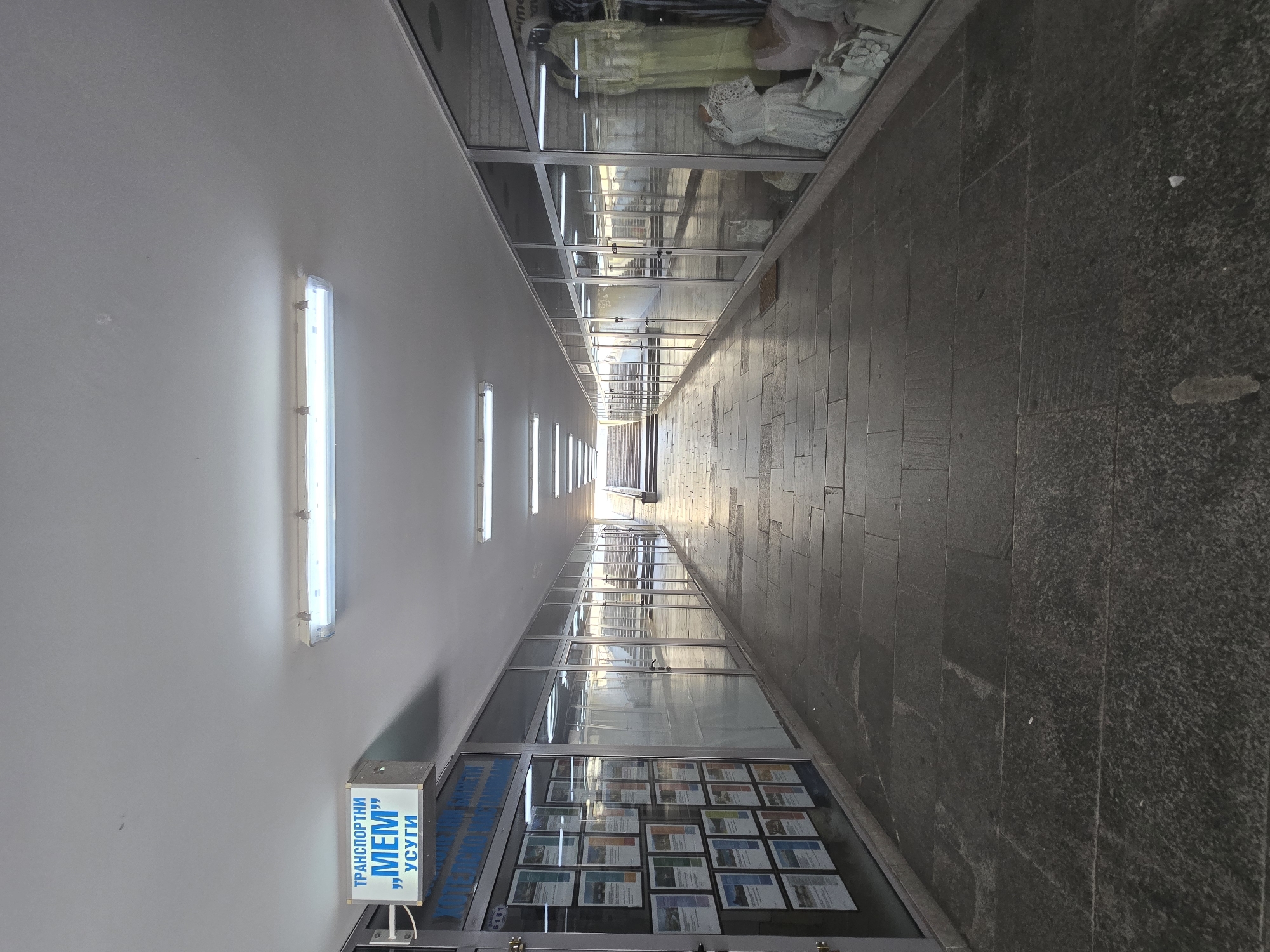 The image shows a long, narrow, covered pedestrian walkway, likely an underpass or a commercial passage, in Plovdiv, Bulgaria. The walkway has shops with glass storefronts on both sides. The floor is paved with large, dark stone slabs. The ceiling is white and illuminated by fluorescent lights.
On the left side, a shop displays numerous informational posters and flyers behind its glass facade. A sign above this shop reads "ТРАНСПОРТНИ „MEM” УСУГИ" which translates to "TRANSPORT „MEM” SERVICES."
On the right side, a shop displays women's clothing and accessories, including dresses and handbags, in its window.
At the far end of the walkway, a set of stairs leads upwards, suggesting an exit or an elevated area. The walkway is empty of people, and the overall atmosphere is subdued, likely due to the lighting and the absence of activity. Sunlight is visible at the very end of the passage, indicating the direction of an exit.