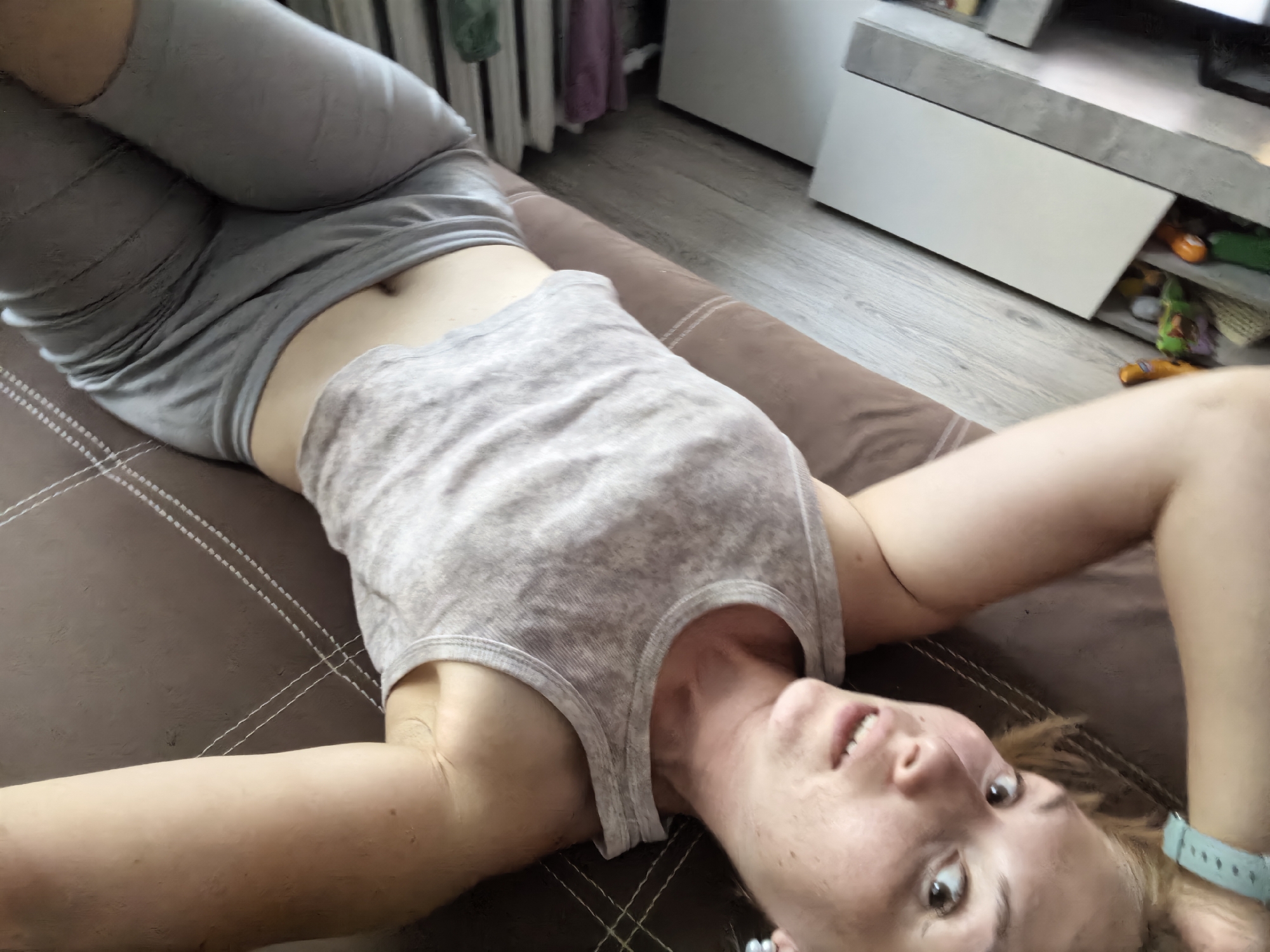 The image shows a person lying on a brown couch. The person is wearing a grey tank top and grey leggings. The person's head is tilted back and their mouth is open, as if they are speaking or laughing. The person is looking at the camera. The background of the image is blurred, but it appears to be a room with furniture. The image is taken from a high angle, looking down at the person. There is no information provided about the location in the city of Sterlitamak, Russia.