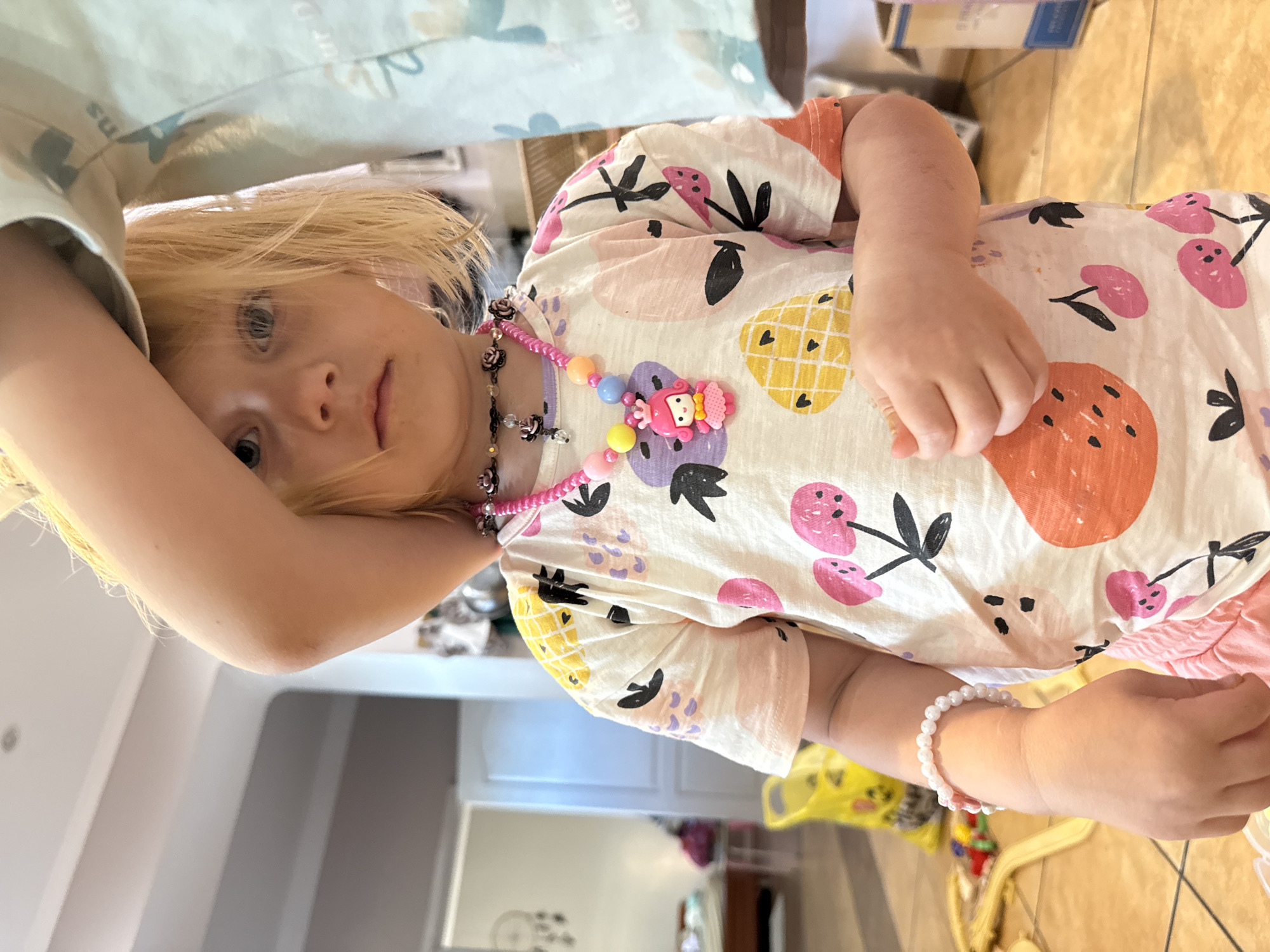A young child with fair skin, blonde hair, and blue eyes is centered in a medium eye-level shot indoors. The child is looking directly at the camera with a neutral expression. Their right forearm is raised, partially obscuring the top of their head. The child is wearing a short-sleeved, light-colored t-shirt patterned with colorful abstract fruit designs including yellow pineapples, pink cherries, and orange citrus slices. Two necklaces are visible: a dark-colored choker with small rose-shaped beads and a clear drop, and a longer pink beaded necklace featuring various pastel-colored spherical beads and a pink cartoon princess charm with a yellow bow. A white beaded bracelet is on the child's left wrist. Pink shorts are visible below the t-shirt.
To the immediate right of the child, a light-colored fabric, possibly part of another person's clothing, is visible, displaying faint light blue patterns and partial text "ns" and "our D".
The background reveals an indoor setting with light-colored tiled flooring and white walls. A doorway leads to another room in the distant left background, where a bed or furniture item is discernible. A decorative dreamcatcher-style object hangs on the wall. A yellow plastic bag or basket containing colorful objects is on the floor to the child's left. A cardboard box is partially visible in the far right background.
Location: Unknown, Unknown.