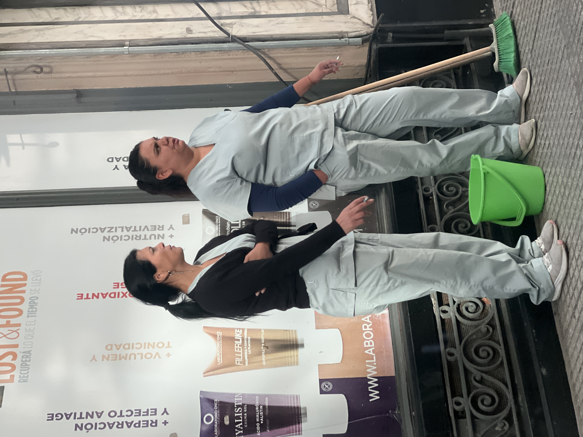Two women in light blue cleaning uniforms stand outside a shop in Buenos Aires, Argentina, smoking cigarettes. One wears a dark sweater over her uniform. A green bucket and a broom are next to them on the sidewalk. The shop's advertising poster is visible, featuring anti-aging creams and the brand name "Laboratorio Once." The poster also includes text in Spanish, like "Lost & Found" and "Recupera lo que el tiempo se llevo." The setting appears to be an urban area.