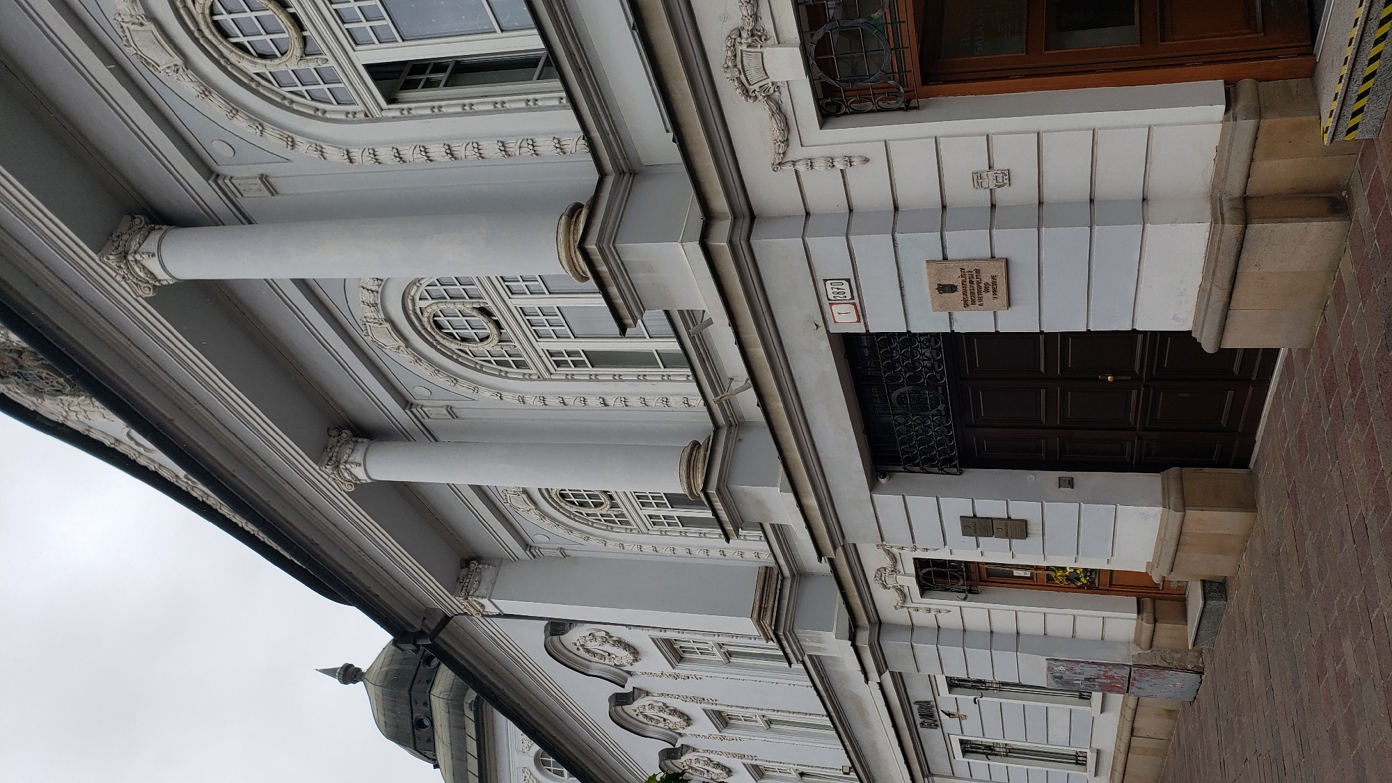 The image shows the exterior of a grand, light blue building with classical architectural elements. The building features large, arched windows with intricate detailing and prominent white columns supporting an ornate entablature. There are several doors visible at ground level, including a large, dark wooden double door. Above the doors are plaques, one with text that appears to be in Slovak. A street sign indicating "1 2870" is mounted on the building. The paving in front of the building is made of red bricks. The sky is overcast. Based on the visible signage and architectural style, this building is likely the J. S. Vrábel Gallery (Galéria J. S. Vrábel) in Prešov, Slovakia, situated on or near Hlavná ulica (Main Street).