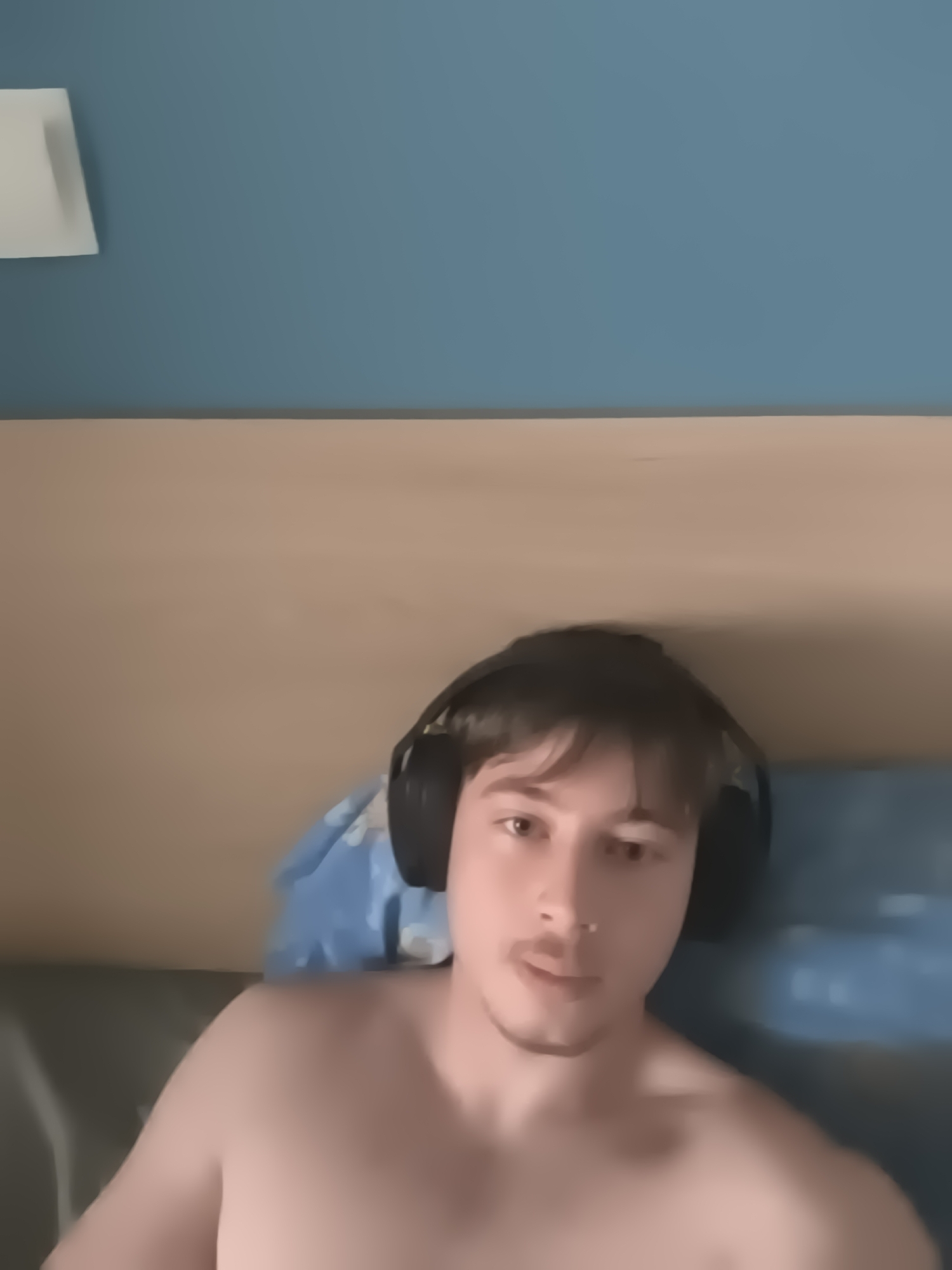 A young man, likely in his late teens or early twenties, is depicted from the chest up in an indoor setting in Sofia, Bulgaria. He has short, dark brown hair, a light mustache, and a faint goatee. He is wearing black over-ear headphones and is shirtless, conveying a casual and relaxed atmosphere. The man looks directly into the camera with a neutral to slightly pensive expression, his lips subtly parted.
The background consists of a two-toned wall. The upper section is a muted, desaturated blue, while the lower part, possibly a bed headboard or wainscoting, is a light tan or beige. To the upper left, a portion of a light-colored, rectangular object, which could be a light fixture or a small shelf, is visible against the blue wall. Behind the man's right shoulder, a crumpled blue fabric object, possibly a blanket or pillow, is discernable. The lighting is soft and diffused, suggesting indoor artificial light or indirect natural light, making the precise time of day indeterminate. There is no visible text in the image. The man's attire and headphones suggest he might be engaged in activities such as gaming, listening to music, or participating in a video call.