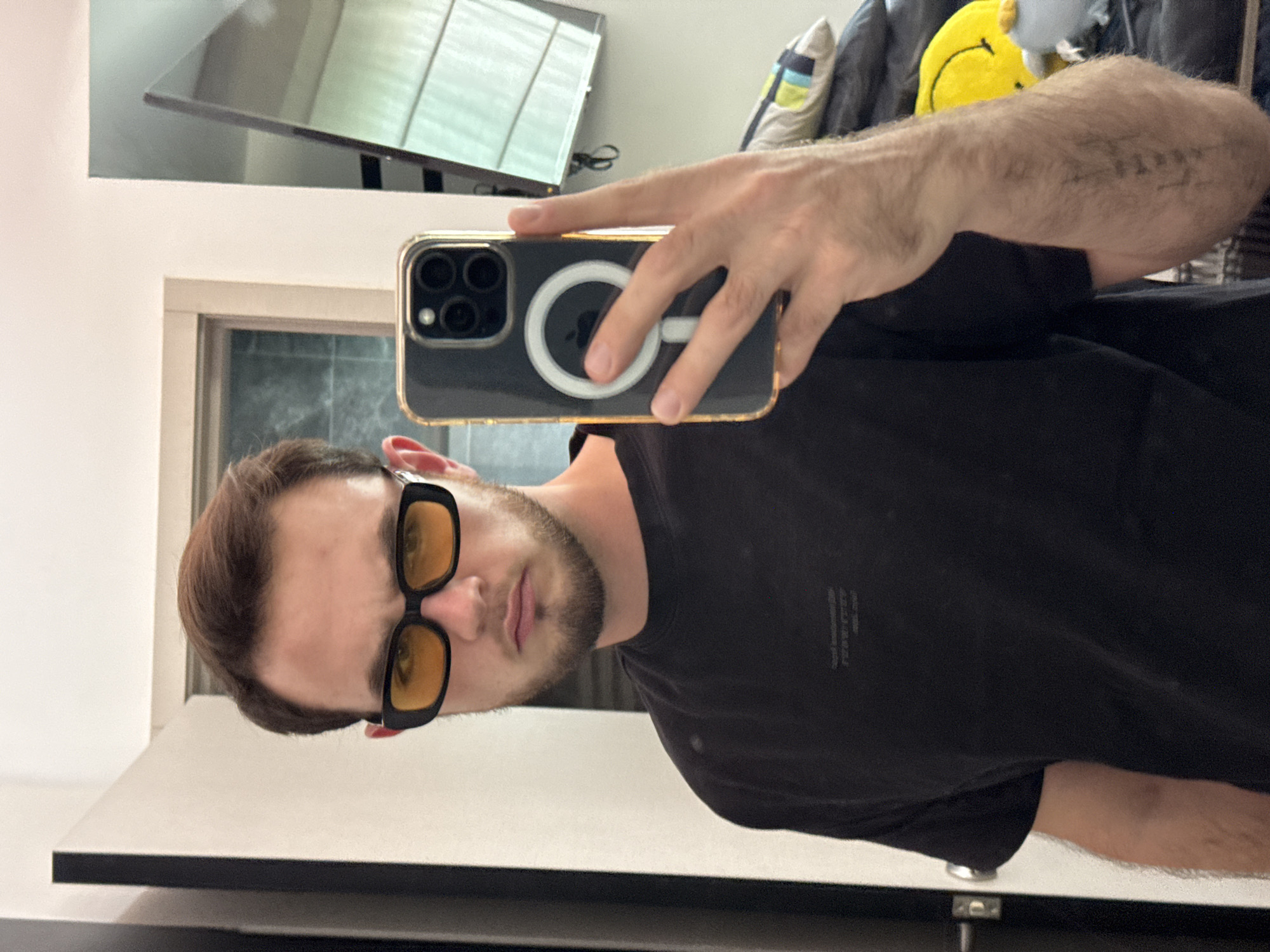 A man takes a selfie in a bathroom mirror. He wears yellow-tinted glasses and a black t-shirt. He is holding a black iPhone with a clear case, MagSafe, and gold trim. A TV is mounted above the mirror. The background behind the man shows a blurred scene with a pillow with a smiley face on it. The location of San Bernardino Tlaxcalancingo, Mexico, cannot be determined from the image.