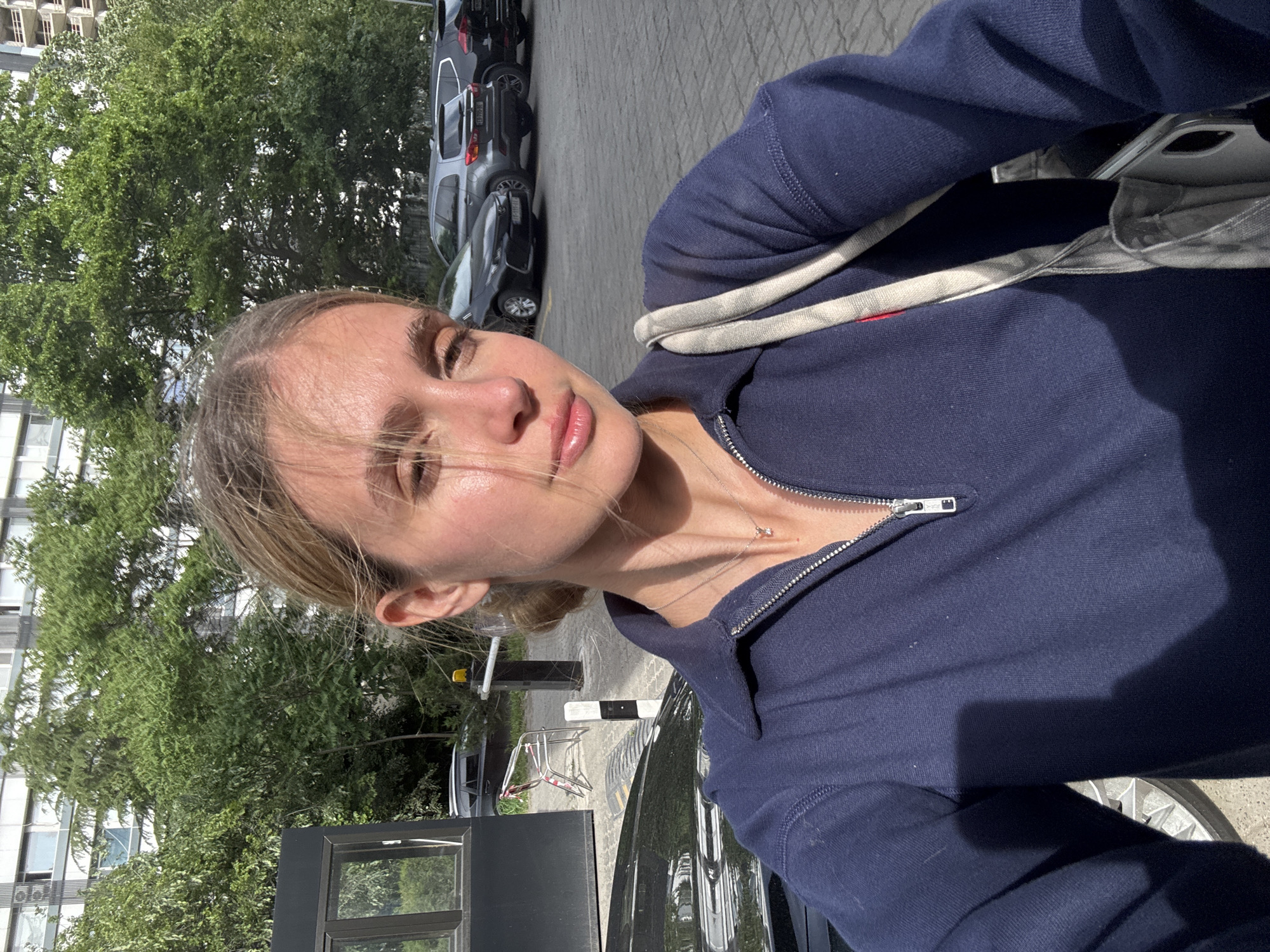 A selfie of a fair-skinned woman with light blonde hair pulled back, wearing a navy blue zip-up sweater and a light-colored bag with a striped strap, taken outside in daylight. The background includes an urban landscape with trees, a multi-story building, cars parked on a paved surface, and a security booth. The location is plausibly Tbilisi, Georgia, based on architectural styles and common urban features in the area. The woman is looking directly at the camera, her sweater zipper is partially down, revealing a necklace. A dark-colored car is visible on the right edge of the frame.