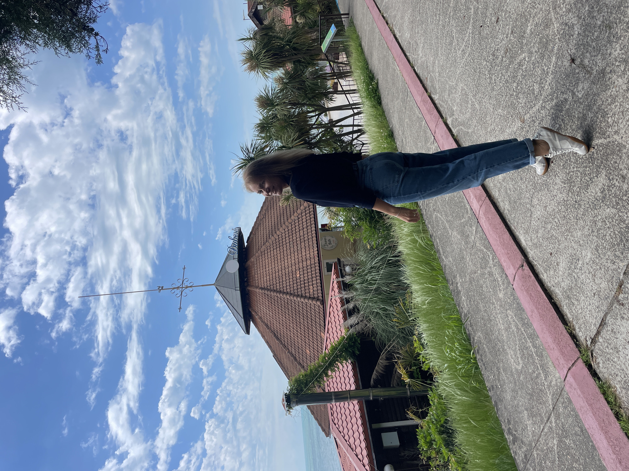 A photograph shows a woman standing outside a building in Sochi, Russia. The woman has long blonde hair and is wearing a dark top and blue jeans. She is standing on a sidewalk with a red curb. Behind her is a building with a red tile roof and a weather vane on top. The sky is blue with white clouds. Palm trees are visible in the background. The sea is visible between the building and foreground grass.
