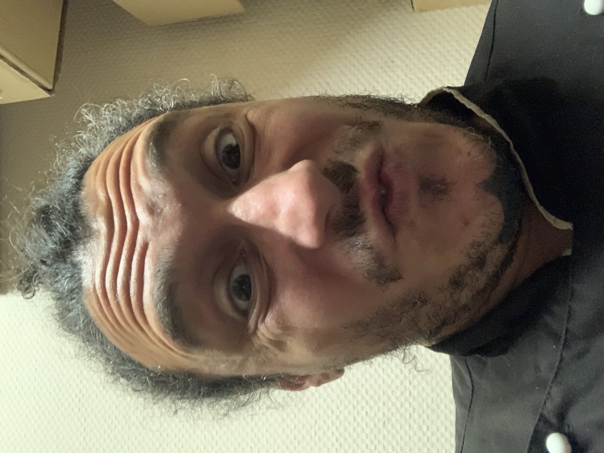 A close-up, low-angle selfie shows a man with curly, dark hair pulled into a bun on top of his head. He has a furrowed brow, wide eyes, and his lips are pursed. He has a mustache and goatee. He is wearing a black chef's jacket with white buttons. The background is a textured wall. The image appears to have been taken indoors, possibly in a restaurant kitchen. The man's expression is one of playful surprise or curiosity. The lighting is dim, casting shadows on his face. There are no indications of the city of Saint Petersburg, Russia, or any specific location within it.