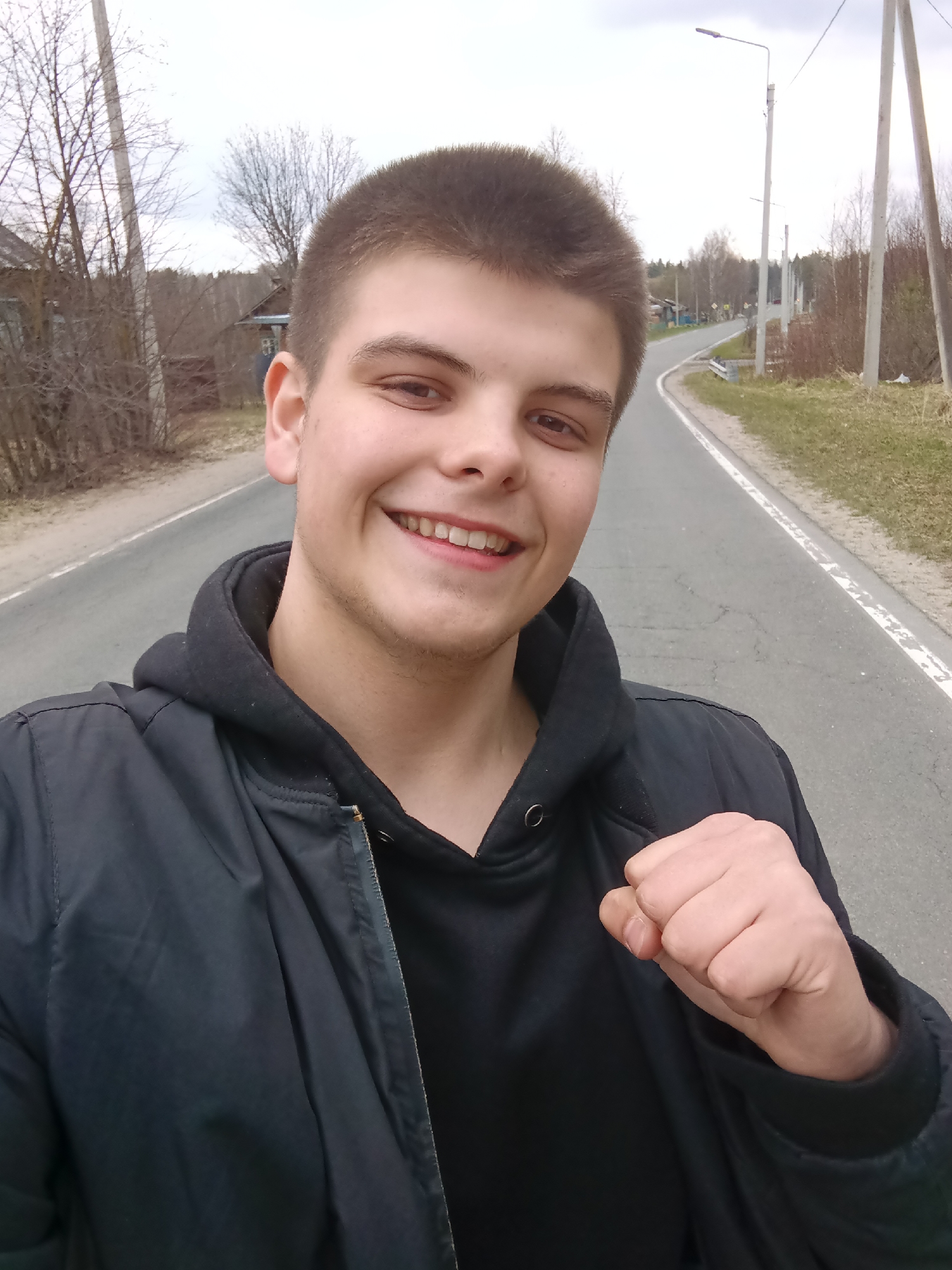 A young man is standing on a rural road in Красный Маяк, Russia, facing the camera. He is smiling and has short, cropped brown hair. He is wearing a black hooded sweatshirt under a dark-colored jacket. His right hand is clenched into a fist near his chest.
The road stretches into the distance, curving to the right. A white line marks the edge of the road. On either side of the road, there is grassy vegetation and trees. Utility poles are visible along the road. In the background, there are houses and more trees, indicating a residential area. The sky is overcast.