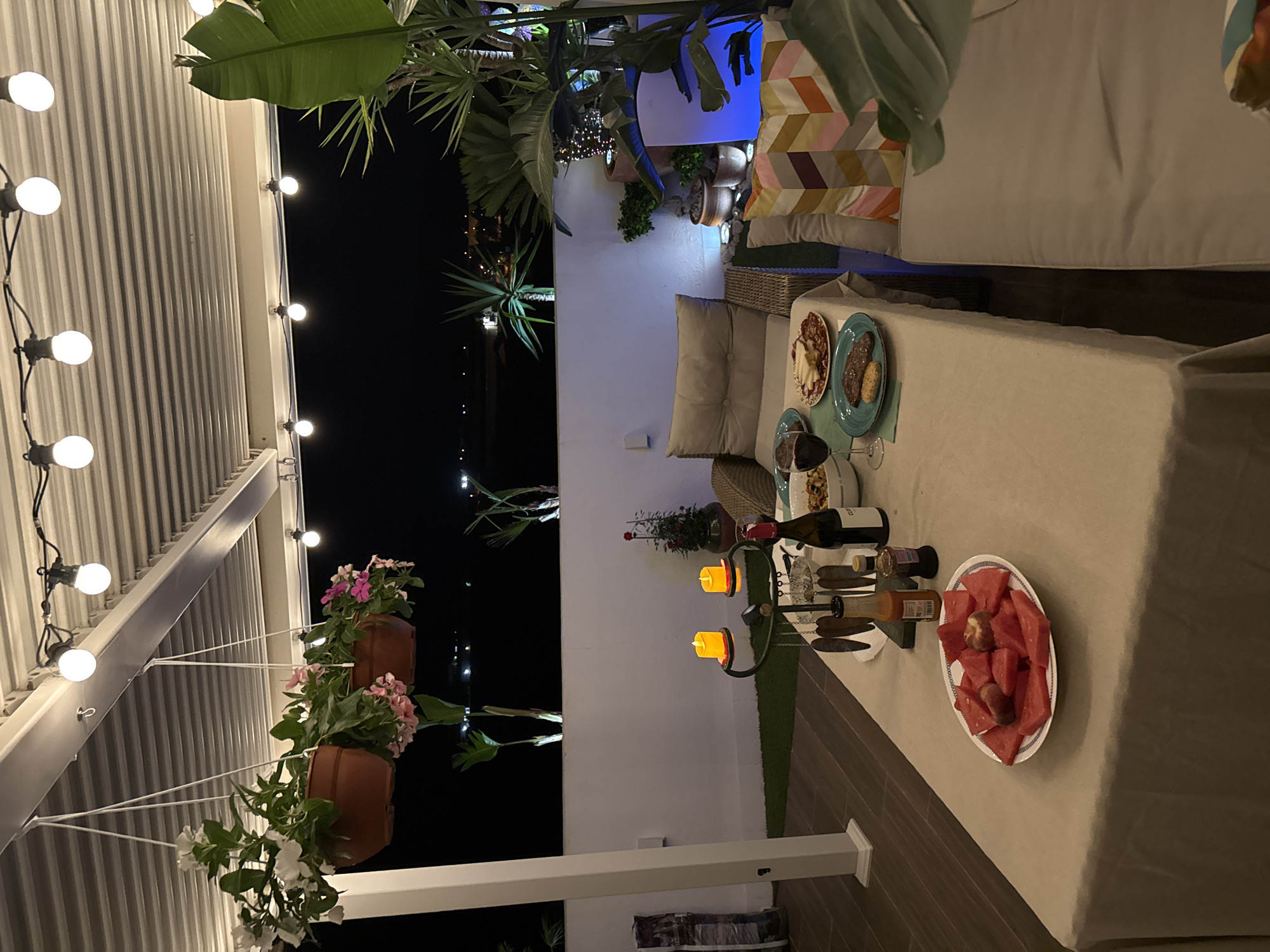 This is a night time, eye-level, wide shot of a covered outdoor dining area on a rooftop terrace in Vallada, Spain. The scene depicts a beautifully set table for a romantic dinner.
In the foreground, a long dining table is draped with a light beige tablecloth. On the table, a plate of watermelon slices with some peaches, a bottle of red wine, a bowl of pasta or salad, and two plates of food are visible. One plate holds what appears to be charcuterie and cheese, while another has a steak and a bread roll. There are also two lit yellow candles on a decorative stand, and various bottles of condiments and utensils.
Behind the table, a wicker chair with cushions is positioned, suggesting comfortable seating. Further back, a white railing separates the terrace from the night sky, where distant city lights can be seen. Lush green plants and trees are strategically placed around the terrace, some in pots and others planted in the ground. Hanging flower baskets with white and pink blooms are suspended from the covered ceiling, adding a touch of color and charm. String lights with round bulbs are draped across the ceiling, casting a warm glow over the entire area. The floor is covered with wooden decking and a patch of artificial green turf. A white pillar on the left side of the image anchors the covered structure. The overall ambiance is one of intimacy and celebration.
