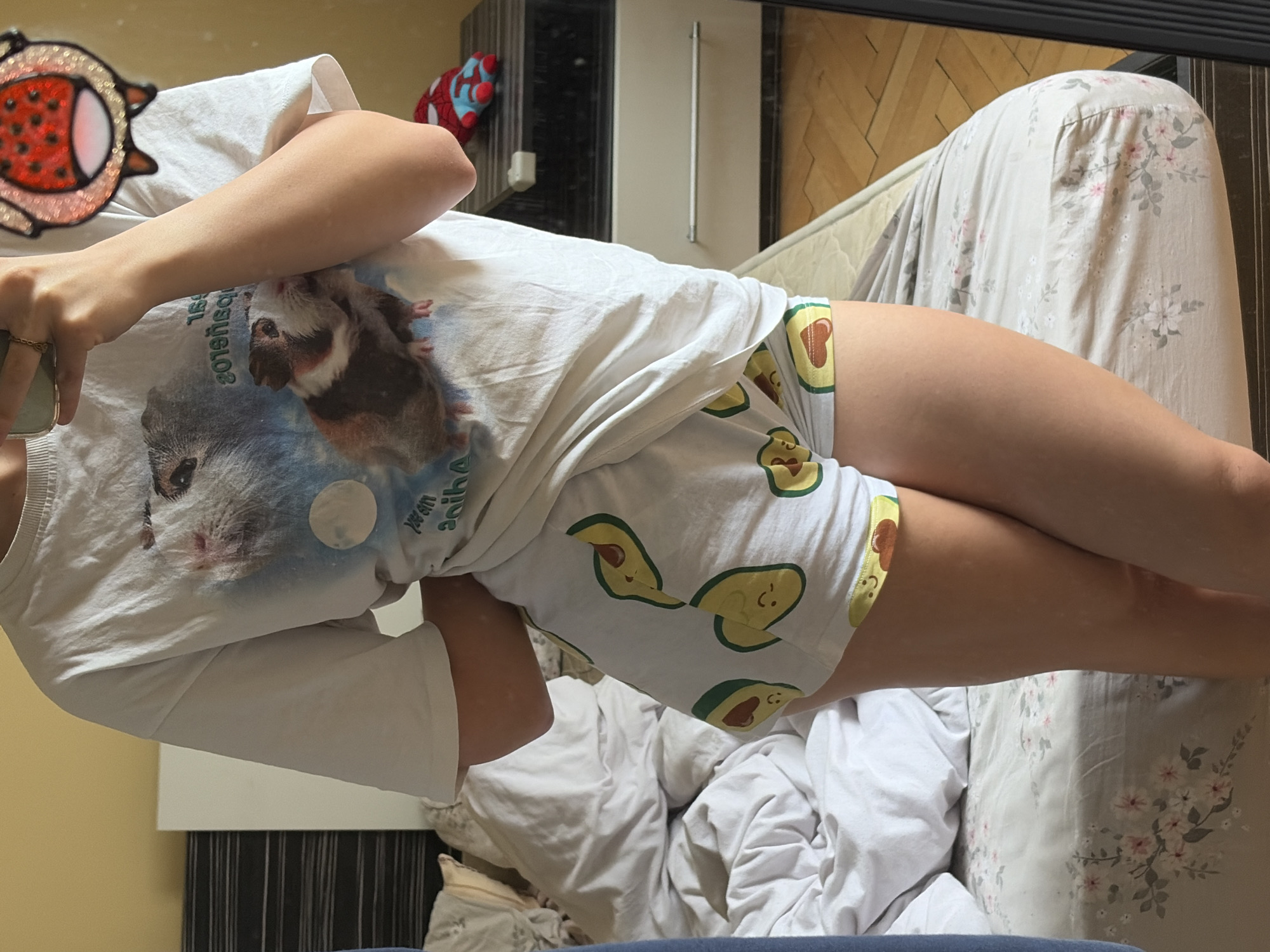 A young person, viewed from the waist down, stands in a bedroom. They are wearing a white t-shirt with a graphic of guinea pigs and shorts printed with avocados. The person is holding a phone in their right hand, taking a selfie in a mirror. The room appears to be a bedroom with a bed, nightstand, and a dresser. The bed is unmade, with white sheets and a floral patterned duvet. On the nightstand, a small stuffed toy resembling Spider-Man is visible. The floor is made of wooden planks.