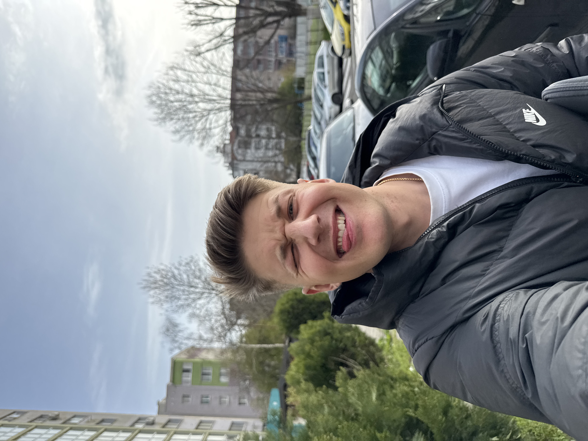 A man is posing for a selfie outdoors. He is wearing a black Nike puffer jacket over a white t-shirt, and a gold chain is visible around his neck. He is smiling broadly with his tongue partially out and his eyes squinted.
Behind him are several parked cars, and in the background, there are buildings and trees. The buildings appear to be residential apartments. The sky is overcast. The trees are leafless, suggesting it might be early spring or late autumn. The location appears to be in Burgas, Bulgaria.