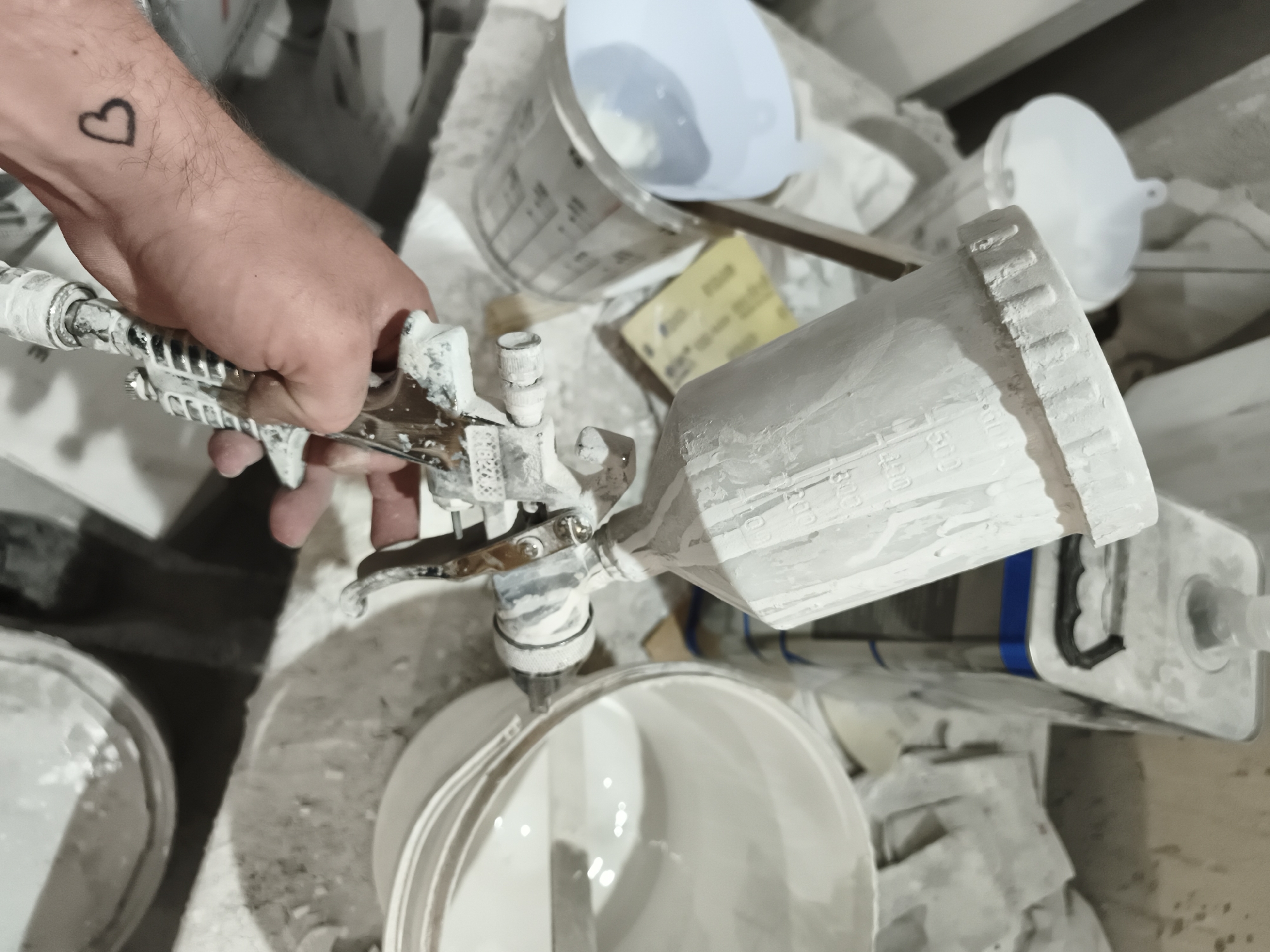 A person's hand, bearing a heart tattoo on the wrist, holds a paint sprayer covered in white paint. The background includes buckets, containers, and tools, also covered in white paint, suggesting a painting or refinishing project. The location is Unknown, Unknown.