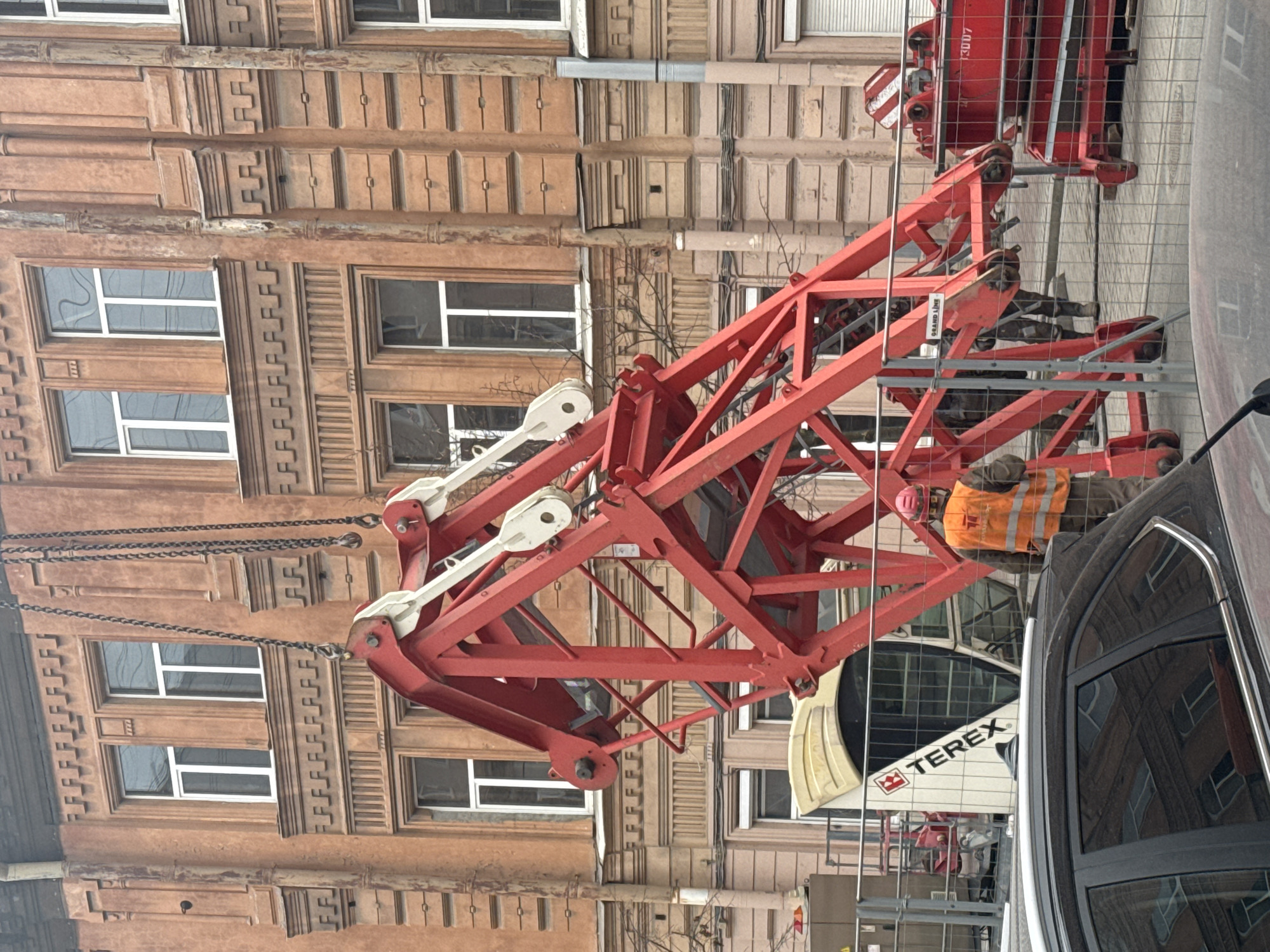 This media file captures an urban construction scene in Saint Petersburg, Russia, featuring a large red crane in the foreground against the backdrop of an ornate, older building. The image appears to be taken during daytime under an overcast sky.
The prominent subject is a massive red crane, which seems to be either in the process of assembly or disassembly. Its substantial steel framework dominates the central and right portions of the image. Several heavy-duty chains are attached to the crane's components, suggesting ongoing lifting or positioning. Part of the crane's counterweight or engine housing is visible at the top right, bearing the white text "3007" on its red surface. Below, another white component with "TEREX" written in black is visible, likely part of the crane's base or an associated vehicle. A person in an orange high-visibility vest and a pink hard hat is seen standing behind a safety barrier near the crane's base, indicating construction work.
To the right, a black car is partially visible, with its glossy surface reflecting parts of the crane and the building.
The background consists of a multi-story building, characteristic of older European architecture. It features a light brown or tan facade with rich, classical detailing, including decorative cornices, pilasters, and intricate reliefs above and between numerous windows. Many of the windows have white frames, and some reflect the sky or surrounding structures. A dark drainpipe runs vertically down the building's facade.
The scene depicts an active construction or infrastructure project within a historical urban setting, with modern heavy machinery juxtaposed against classic architecture. Another small label "GRAND LINE" is also visible on a white tag attached to the crane structure.
