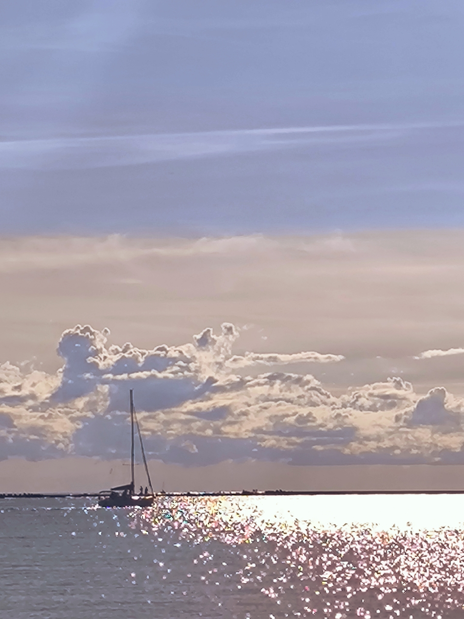 The image depicts a sailboat on the water, viewed from a distance. The sailboat has a tall mast and appears to have several people on board. The water is calm and reflects the sunlight, creating a shimmering effect. In the background, there are clouds in the sky, with hints of purple and pink hues. The overall scene is serene and suggests a peaceful moment on the water. The location is not specified in the image, and therefore, it cannot be identified as Riga, Latvia.