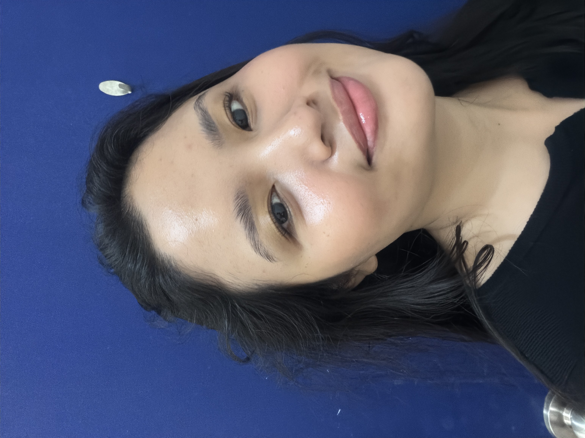 A medium-shot, selfie-style photograph shows a young woman with long, dark hair and light skin. She is smiling, with her head tilted slightly to her left. The woman is wearing a black, ribbed top. Her makeup includes foundation, blush, mascara, and lip gloss. The background is a solid, dark blue wall with a small, white hook visible to the upper right of her head. The lighting suggests an indoor setting. The image does not provide any specific visual cues that would indicate its location within Davao City, Philippines.