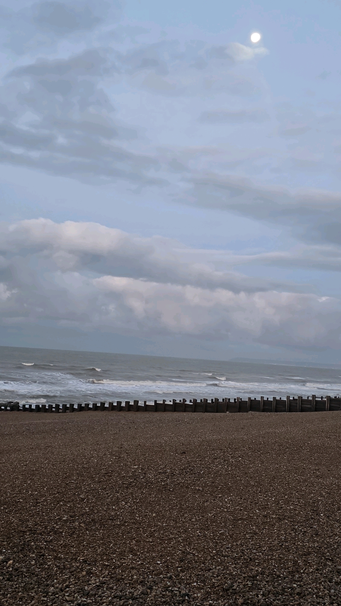 The image depicts a beach scene, likely in St Leonards, United Kingdom, during twilight or an overcast day. The foreground is dominated by a densely packed expanse of pebbles. Beyond the pebble beach, a line of wooden groynes stretches horizontally across the frame, jutting out towards the sea. The sea itself is active, with visible waves breaking and white foam along the shore. The water appears grey and choppy, suggesting a moderate wind. The sky is a prominent feature, filled with a mixture of light and dark grey clouds, with a brighter, paler area in the upper right quadrant where a full or nearly full moon is visible, partially obscured by wispy clouds. There are no people or animals visible in the image. The overall atmosphere is somewhat somber and natural, emphasizing the elements of sea, sky, and shore.