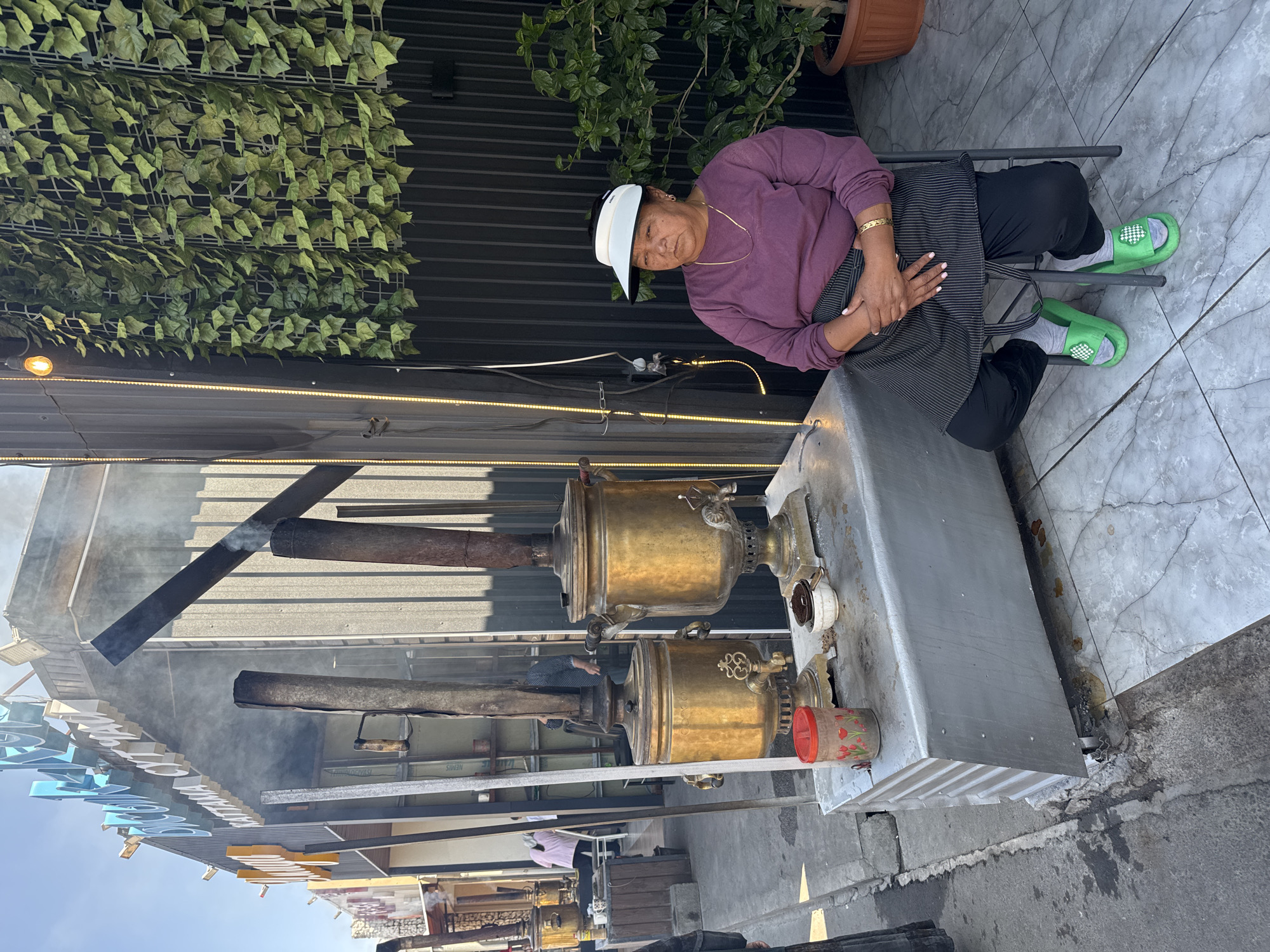 A woman sits on a stool in front of a stand with two large, brass samovars, which are heating devices used for boiling water or brewing tea. The samovars are ornate and appear to be old, with smoke rising from their chimneys. The woman is wearing a purple long-sleeved shirt, black pants, a striped apron, a white visor, and green slippers. She is looking off to the side with her hands clasped in her lap.
Behind the samovars, a building with a gray corrugated metal facade is visible. A sign with blue lettering in Cyrillic script hangs above the entrance. There is also a wall covered in artificial green ivy to the right of the samovars. A potted plant with green leaves is situated next to the ivy wall. The ground is tiled with a gray and white marble pattern.
The overall scene suggests an outdoor tea or food stall, possibly in a marketplace or along a street. The presence of the samovars and the woman attending them indicates that this is likely a functioning establishment. The location is identified as Sasyk-Bulak, Kyrgyzstan.